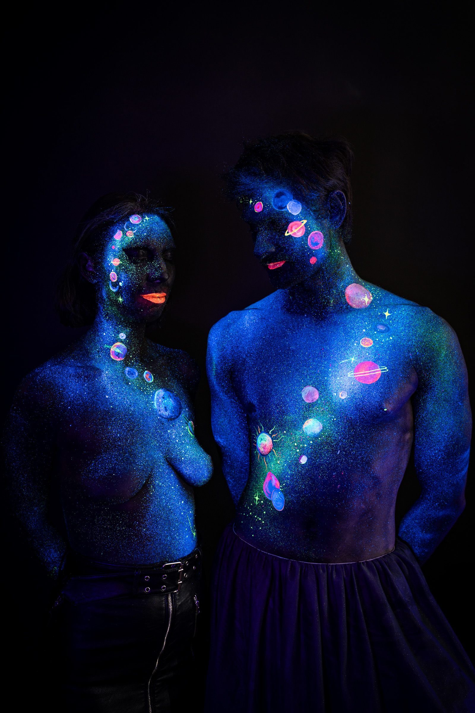 Alessia and Axell stand close together, she looking up toward him while he looks downward toward her. The design is a space themed bodypaint of glowing planets, rings and stars in pink, orange and purple over blue glitter. Bodypaint by Elad Itzkin, Charlotte Bobineau, Sage Brewer. Photographed by Elad Itzkin Photography under UV/blacklight in his studio in Gesundbrunnen, Berlin.