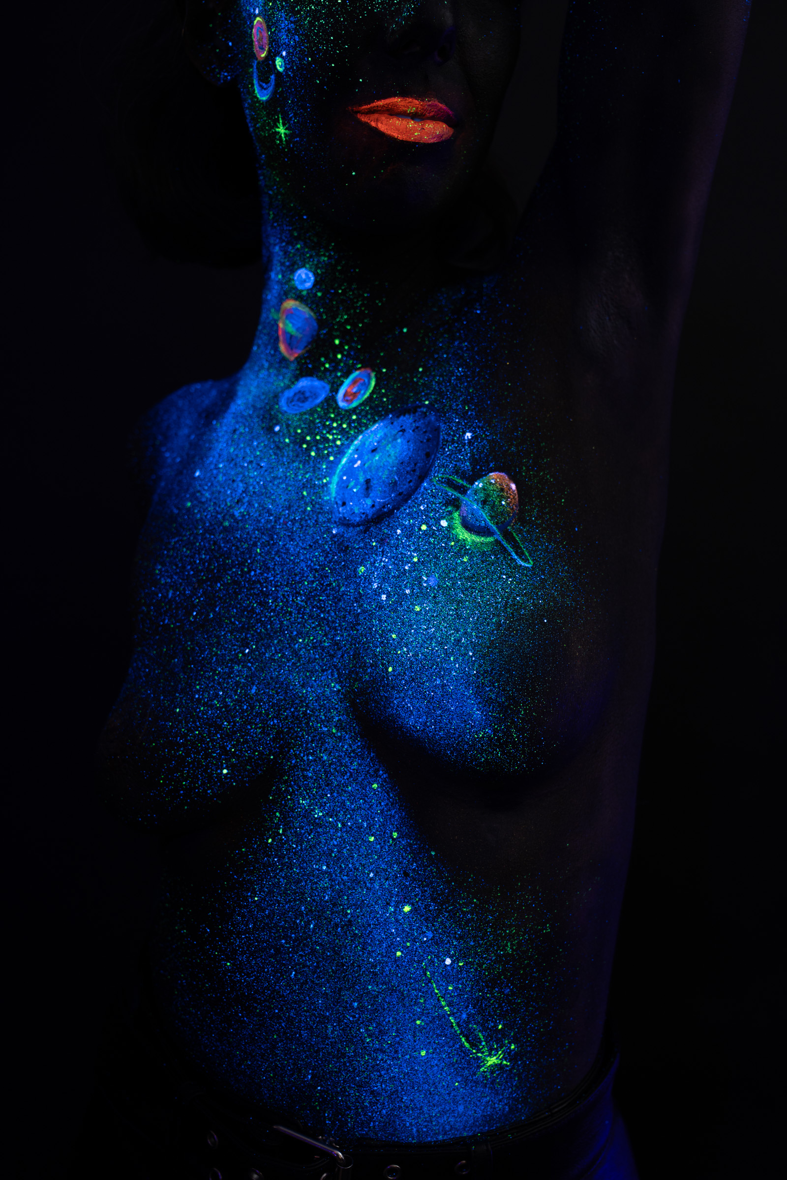 In this close crop, Alessia stands in profile with her neck and chest visible. Blue UV glitter and small orange, pink, and blue planet motifs form a cosmic themed design across her skin. Bodypaint by Elad Itzkin, Charlotte Bobineau, Sage Brewer. Photographed by Elad Itzkin Photography under UV/blacklight in his studio in Gesundbrunnen, Berlin.