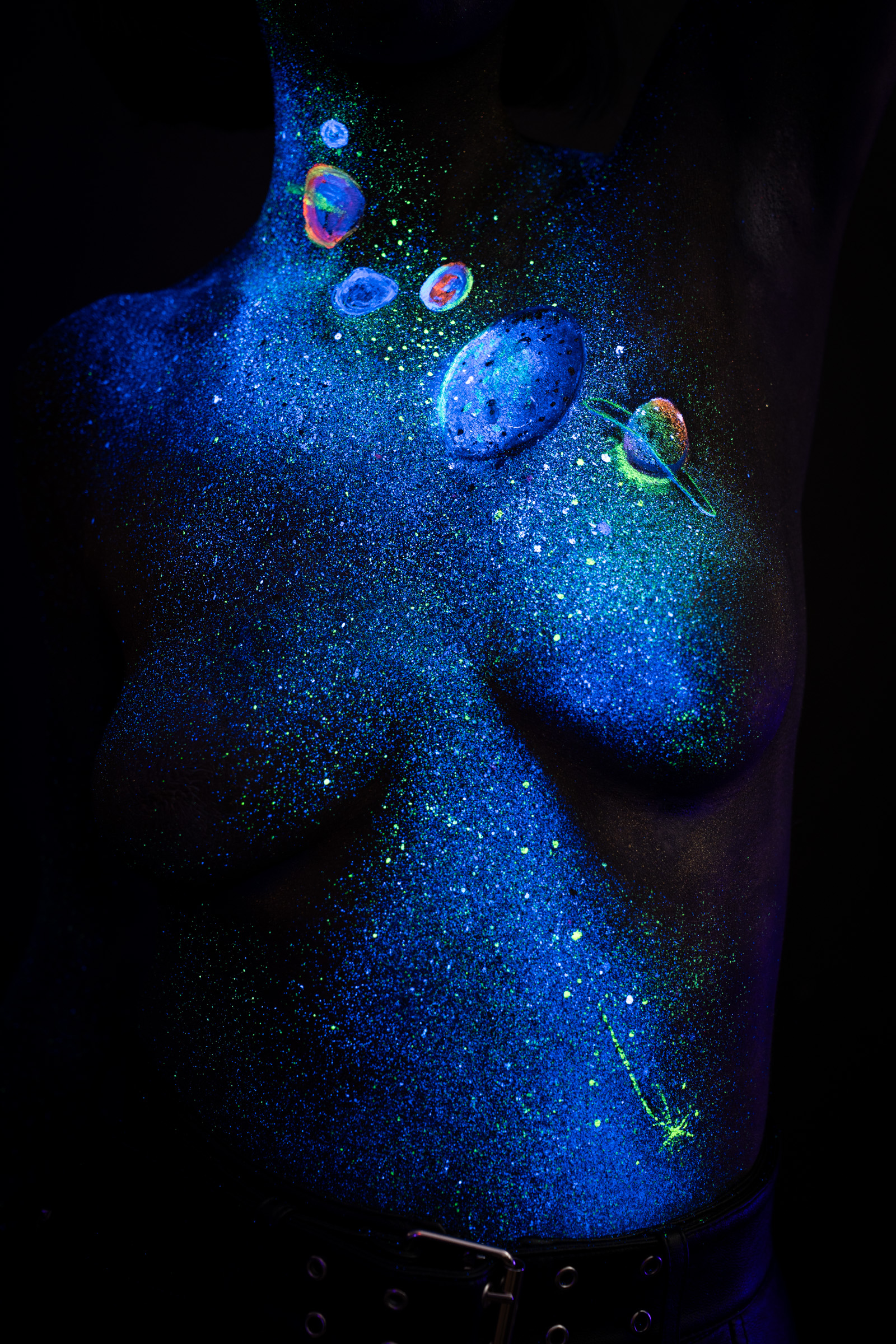A close crop of Alessia's torso shows blue UV glitter with a cluster of small multicolored planet circles and a green shooting star, part of a cosmic themed bodypaint design. Bodypaint by Elad Itzkin, Charlotte Bobineau, Sage Brewer. Photographed by Elad Itzkin Photography under UV/blacklight in his studio in Gesundbrunnen, Berlin.