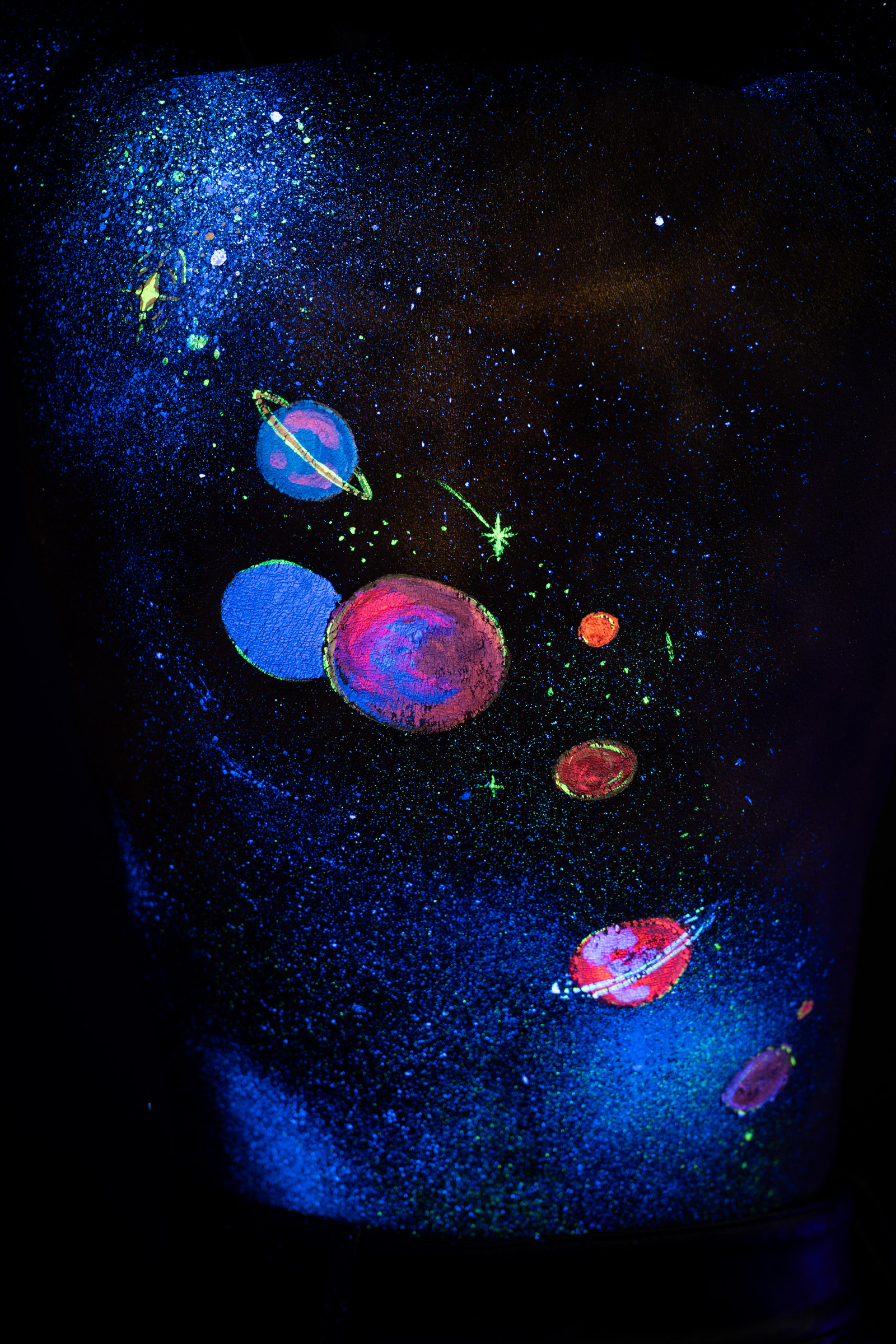 This close crop shows Alessia's back turned to the camera, painted with blue UV glitter and a cluster of small colored planet motifs alongside a green comet streak. Bodypaint by Elad Itzkin, Charlotte Bobineau, Sage Brewer. Photographed by Elad Itzkin Photography under UV/blacklight in his studio in Gesundbrunnen, Berlin.