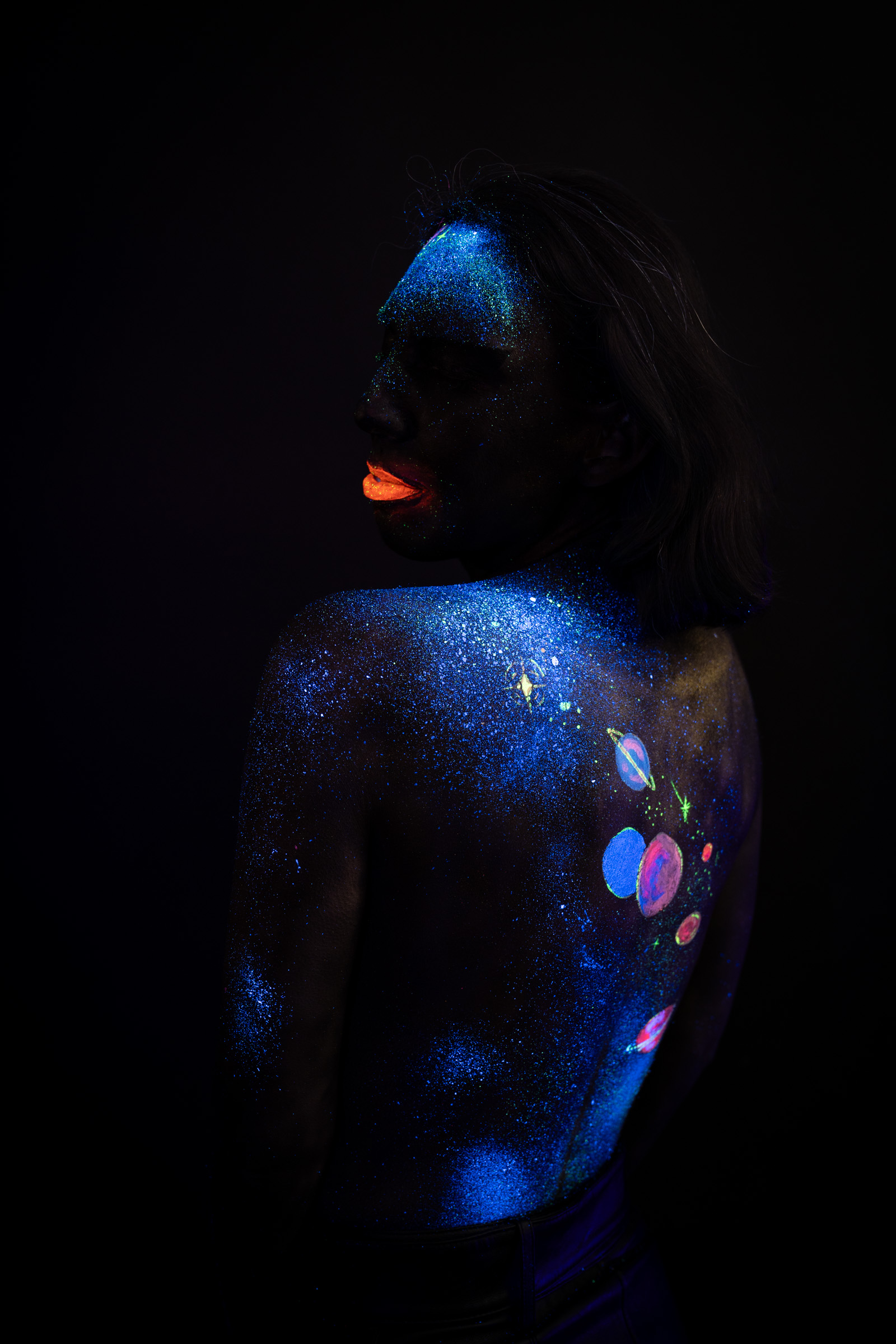 Alessia stands with her back to the camera, head turned to look over her shoulder. A cosmic themed design of blue UV glitter and small planet circles covers her upper back. Bodypaint by Elad Itzkin, Charlotte Bobineau, Sage Brewer. Photographed by Elad Itzkin Photography under UV/blacklight in his studio in Gesundbrunnen, Berlin.