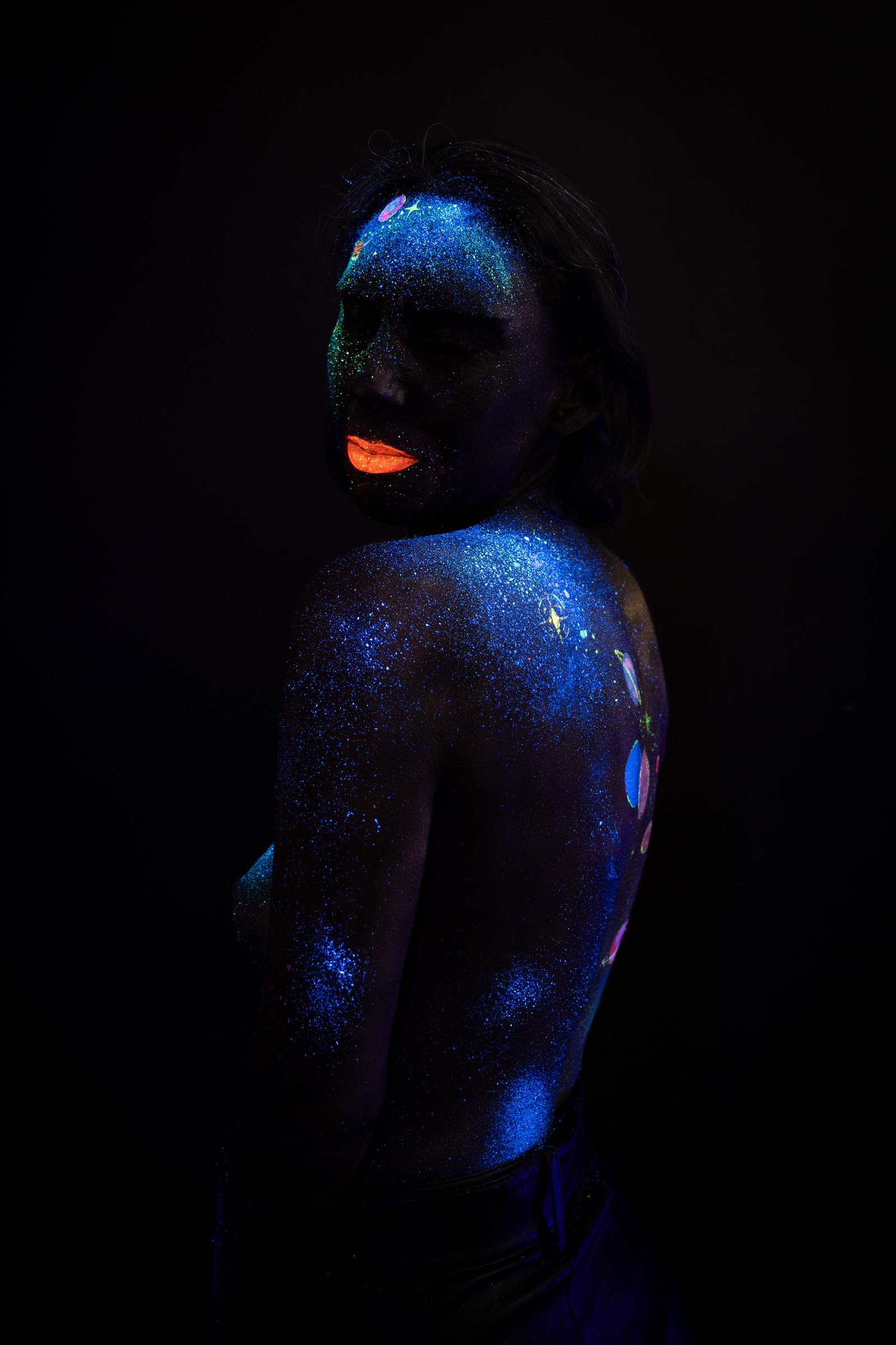 Alessia stands facing forward with her head tilted back. Blue UV glitter and a chain of small colored planet motifs run from her face down her chest in a space themed design. Bodypaint by Elad Itzkin, Charlotte Bobineau, Sage Brewer. Photographed by Elad Itzkin Photography under UV/blacklight in his studio in Gesundbrunnen, Berlin.
