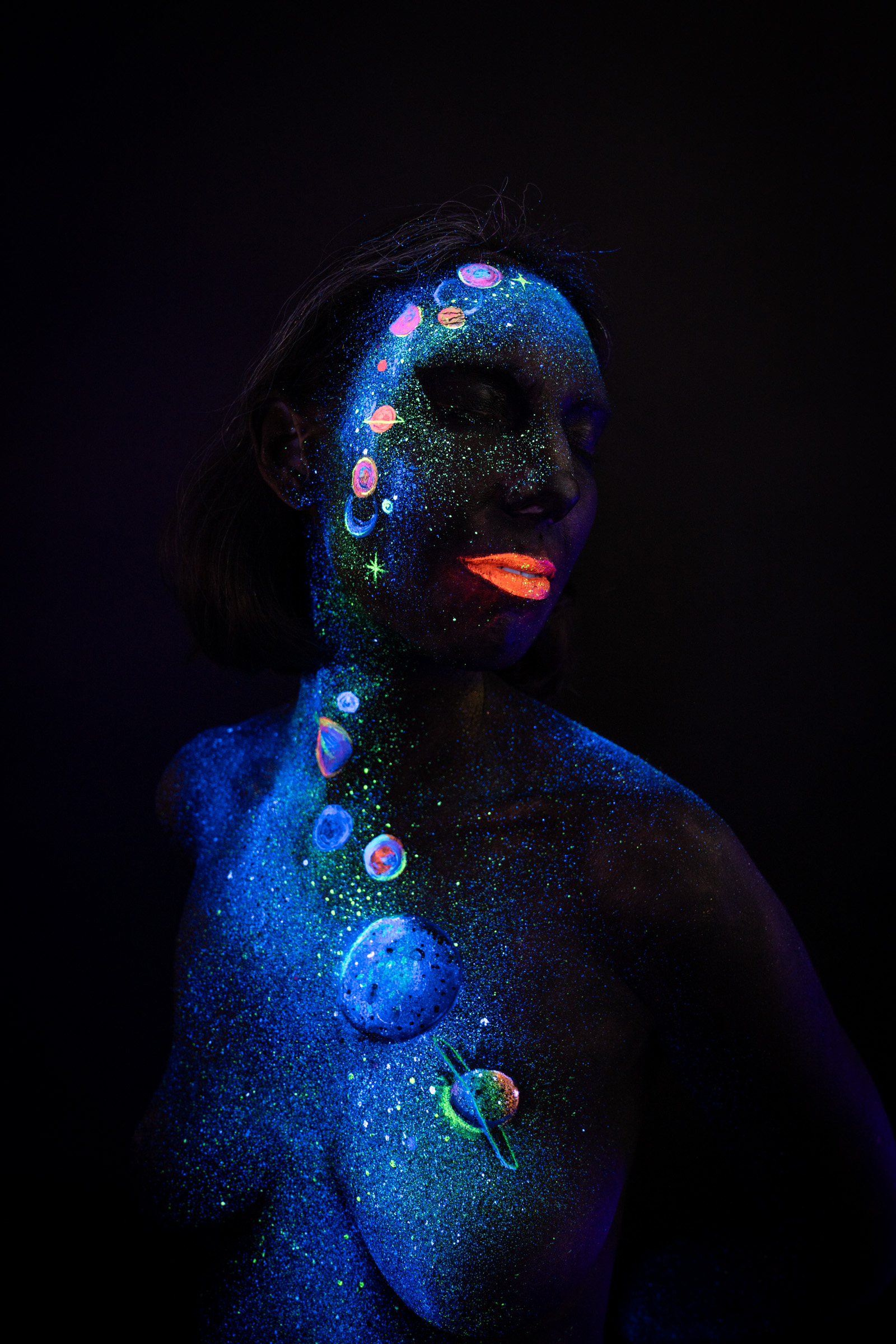 Alessia stands facing the camera at a slight angle. Her face, neck, and chest are decorated with blue UV glitter and small multicolored planet motifs in a cosmic themed design. Bodypaint by Elad Itzkin, Charlotte Bobineau, Sage Brewer. Photographed by Elad Itzkin Photography under UV/blacklight in his studio in Gesundbrunnen, Berlin.