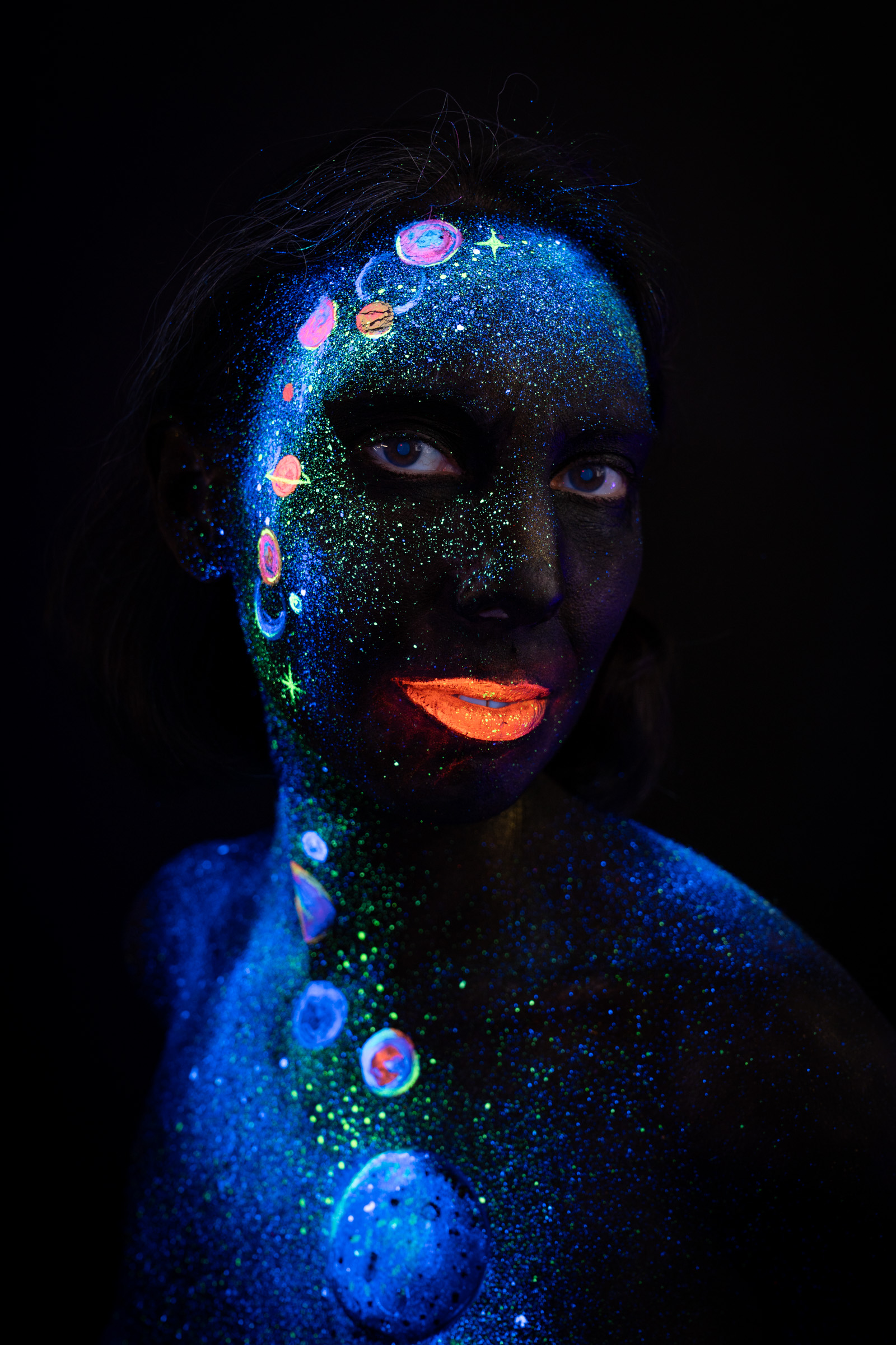 Alessia stands facing the camera directly. A space themed bodypaint design of blue UV glitter with small pastel planet circles extends from her face down her neck and chest. Bodypaint by Elad Itzkin, Charlotte Bobineau, Sage Brewer. Photographed by Elad Itzkin Photography under UV/blacklight in his studio in Gesundbrunnen, Berlin.