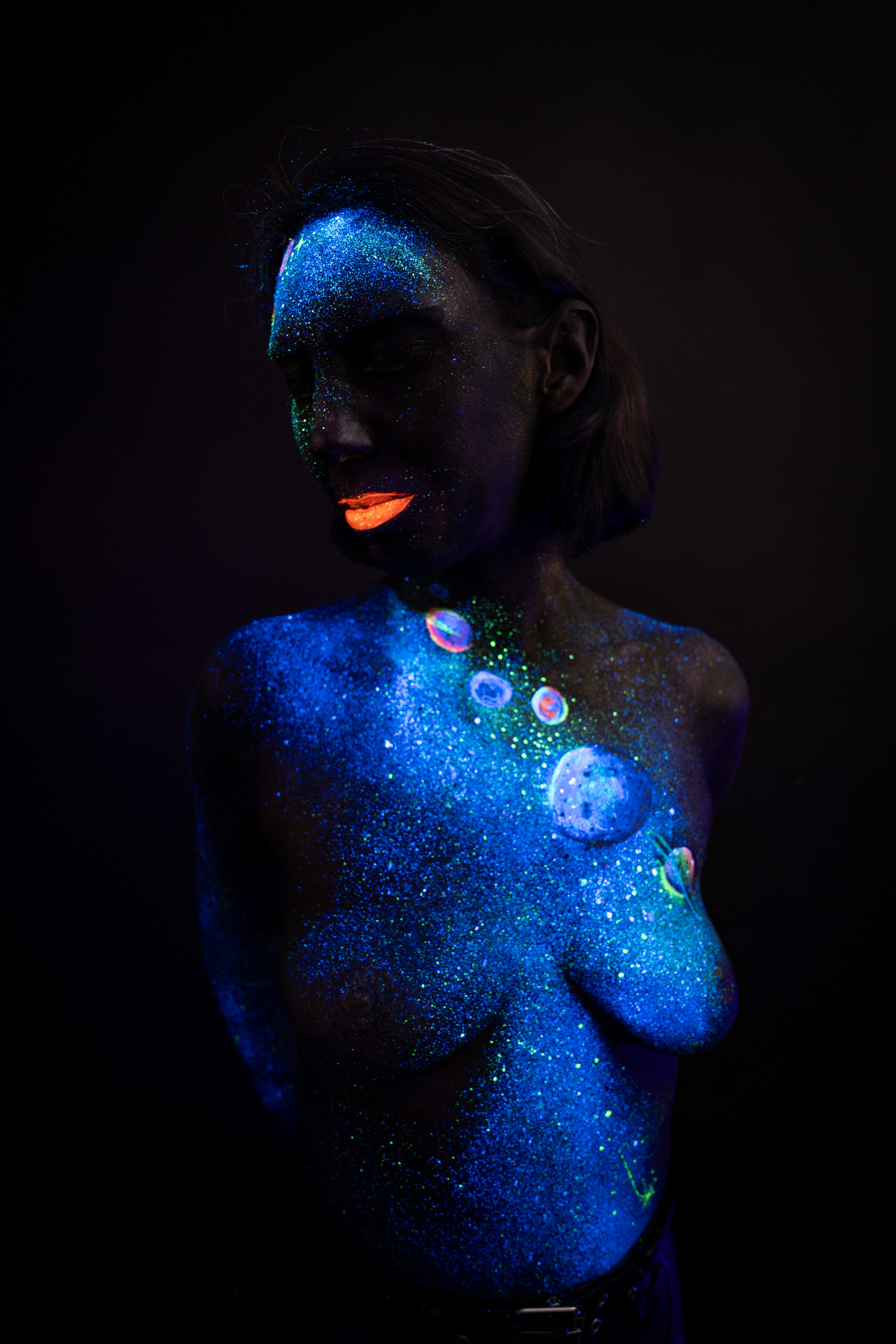 Alessia stands turned to one side with her head angled downward. Blue UV glitter and a cluster of small colored planet motifs cover her chest and shoulder in a cosmic themed design. Bodypaint by Elad Itzkin, Charlotte Bobineau, Sage Brewer. Photographed by Elad Itzkin Photography under UV/blacklight in his studio in Gesundbrunnen, Berlin.