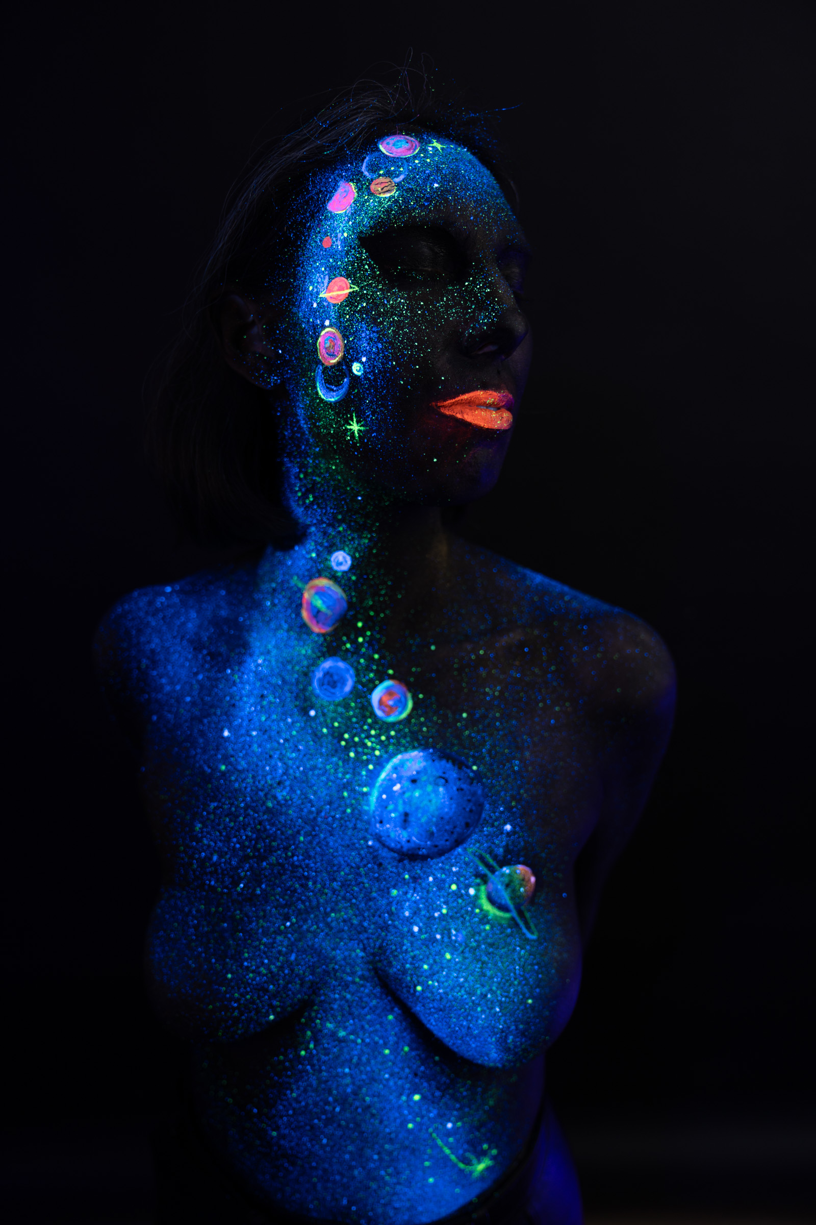 Alessia stands facing the camera at a slight angle, chest exposed. A cosmic themed design of blue UV glitter and pastel planet shapes runs from her face down to her chest. Bodypaint by Elad Itzkin, Charlotte Bobineau, Sage Brewer. Photographed by Elad Itzkin Photography under UV/blacklight in his studio in Gesundbrunnen, Berlin.