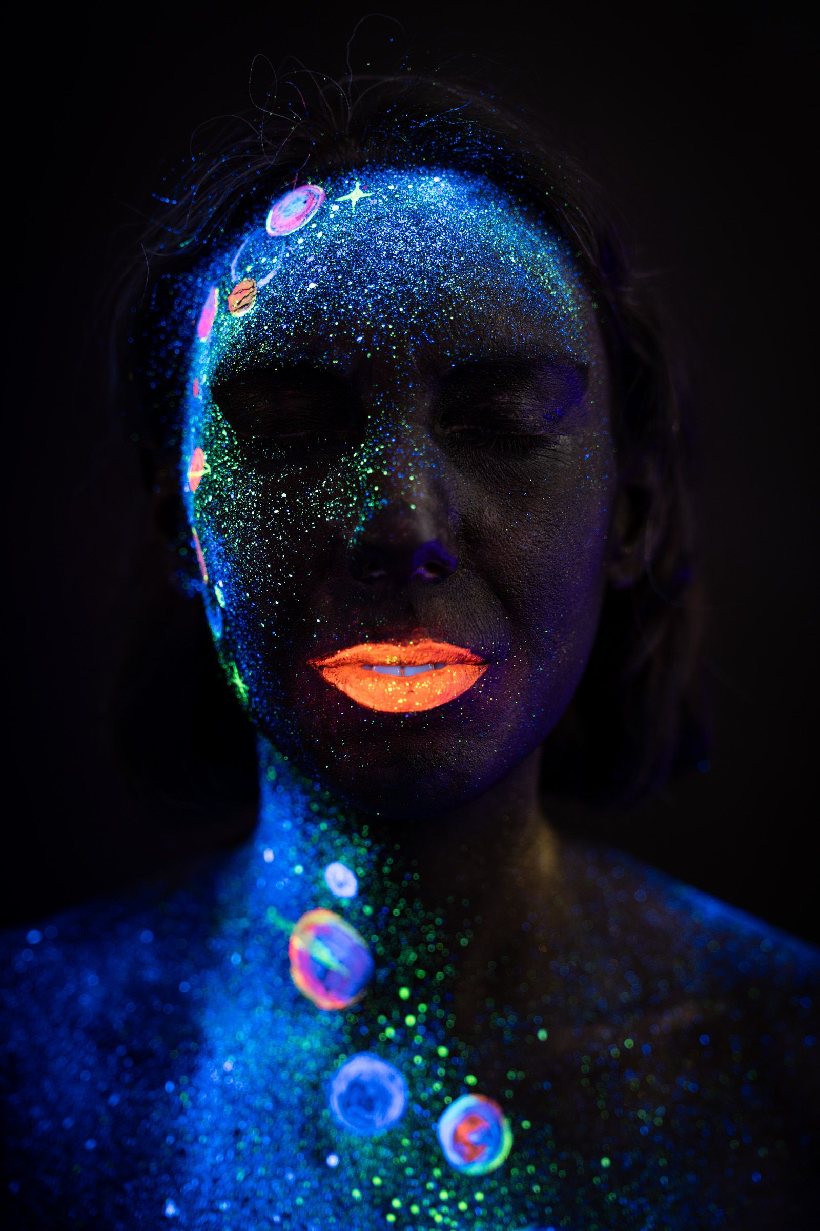 This close-up shows Alessia's face and upper chest decorated with blue UV glitter and small multicolored planet motifs, part of a cosmic themed bodypaint design. Bodypaint by Elad Itzkin, Charlotte Bobineau, Sage Brewer. Photographed by Elad Itzkin Photography under UV/blacklight in his studio in Gesundbrunnen, Berlin.