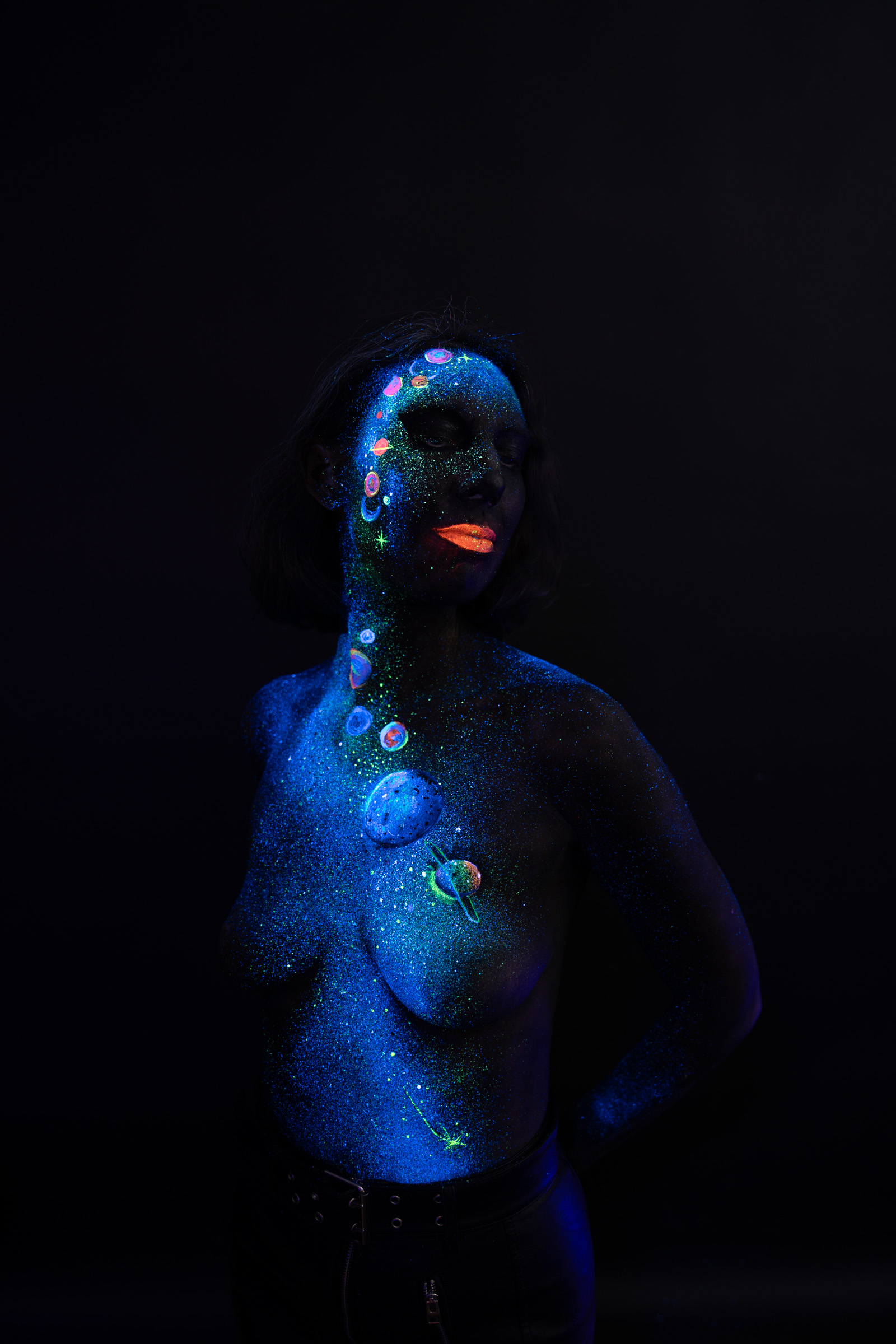 Alessia stands facing the camera. A cosmic themed bodypaint design of blue UV glitter and a cluster of small colored planet motifs covers her chest. Bodypaint by Elad Itzkin, Charlotte Bobineau, Sage Brewer. Photographed by Elad Itzkin Photography under UV/blacklight in his studio in Gesundbrunnen, Berlin.