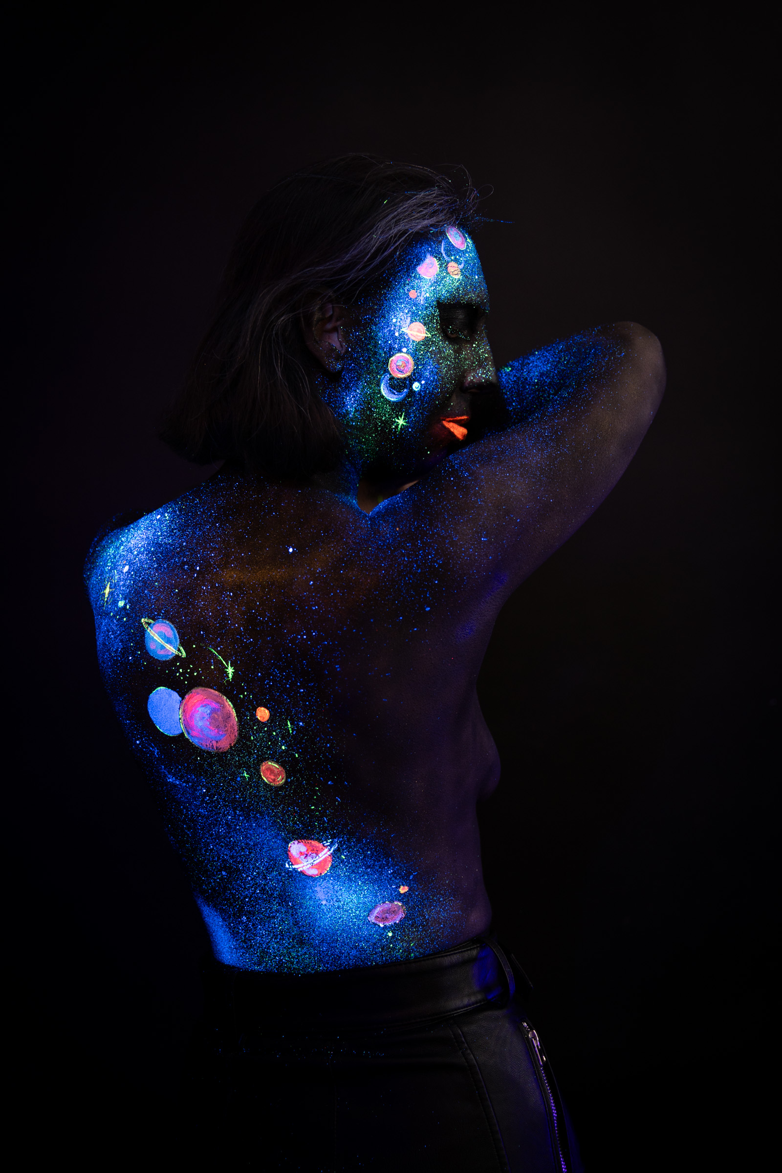 Alessia stands with her back turned, head angled toward the camera. Blue UV glitter and a cluster of small planet circles decorate her back and shoulder in a cosmic themed design. Bodypaint by Elad Itzkin, Charlotte Bobineau, Sage Brewer. Photographed by Elad Itzkin Photography under UV/blacklight in his studio in Gesundbrunnen, Berlin.