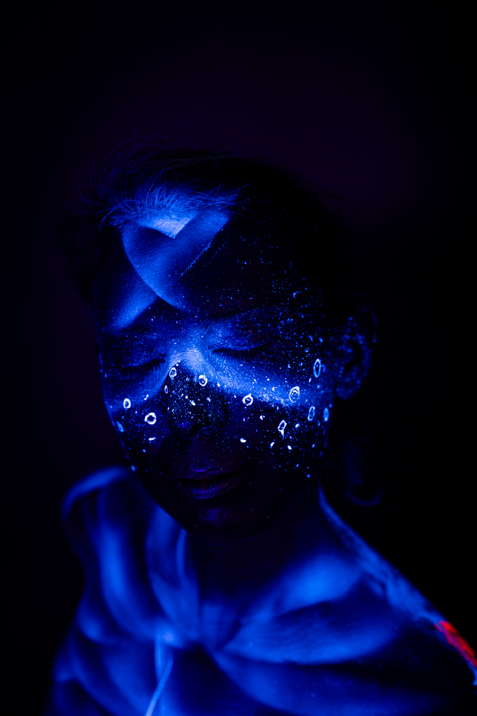 Alessia is shown in a close, three-quarter pose with her eyes closed and head lowered. A blue abstract pattern of dots and droplet shapes spans across her face and upper chest. Bodypaint by Geli, Charlotte Bobineau, Axell, Sage Brewer. Photographed by Elad Itzkin Photography under blacklight in his Gesundbrunnen, Berlin studio.