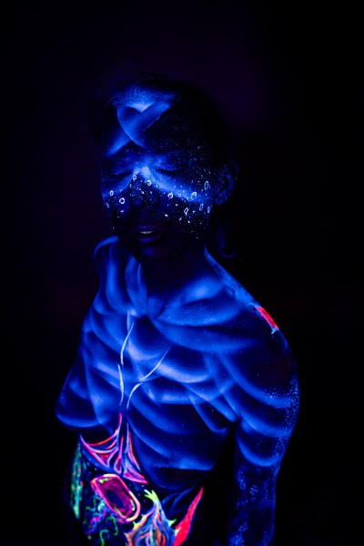 Alessia stands facing the camera with more of her torso visible. Blue linework tracing an abstract pattern covers her chest, with a colorful abstract design at her waist, and dotted blue paint across her face. Bodypaint by Geli, Charlotte Bobineau, Axell, Sage Brewer. Photographed by Elad Itzkin Photography under UV light in his Gesundbrunnen, Berlin studio.