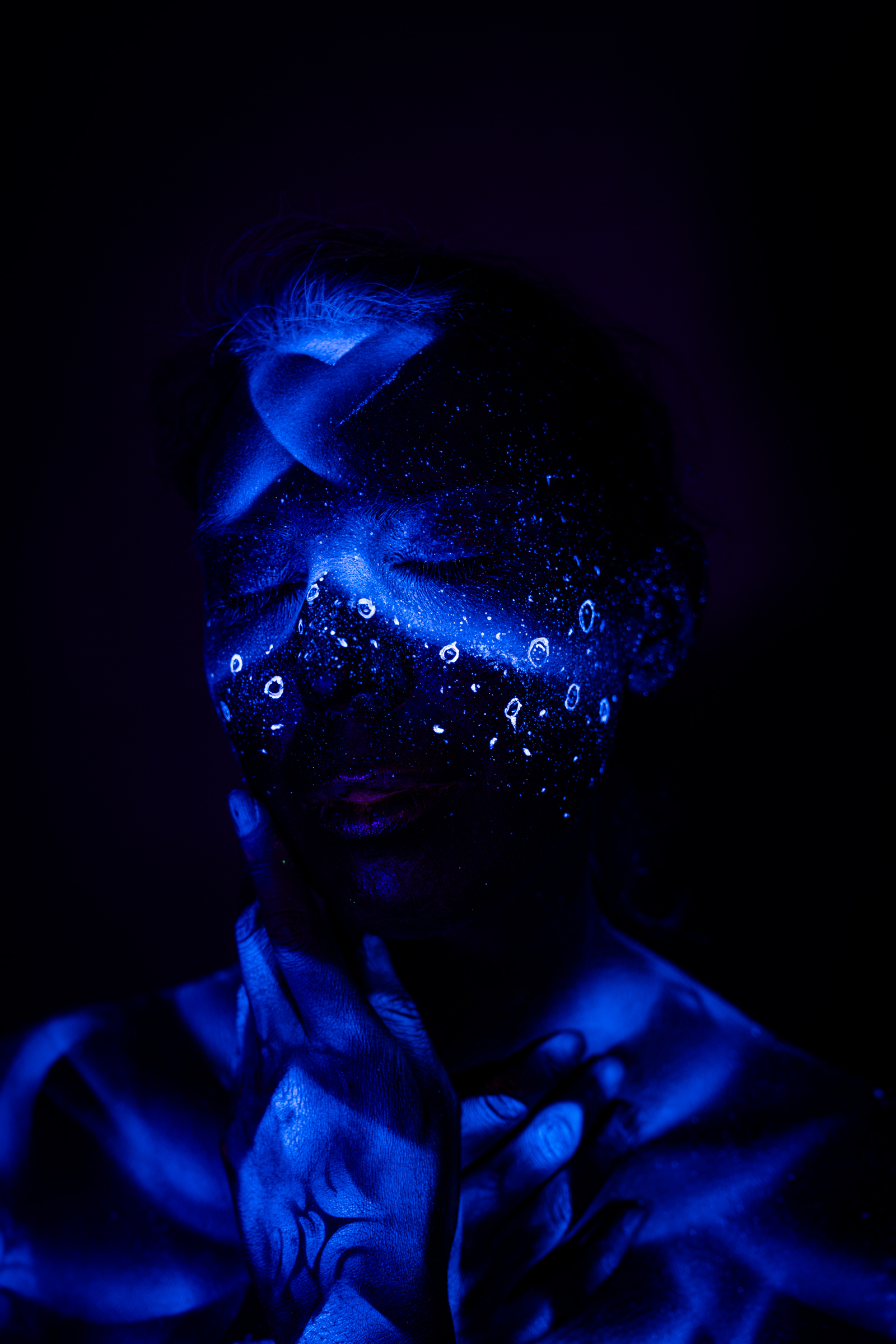 Alessia stands in a close pose with her head lowered. Her face carries a blue abstract dotted design resembling a mask, extending down onto her chest. Bodypaint by Geli, Charlotte Bobineau, Axell, Sage Brewer. Photographed by Elad Itzkin Photography under UV blacklight in his Gesundbrunnen, Berlin studio.