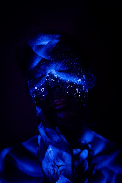 Alessia is photographed in a close, three-quarter pose with her head tilted down. A blue abstract design of dots and droplet shapes covers her face and upper chest. Bodypaint by Geli, Charlotte Bobineau, Axell, Sage Brewer. Photographed by Elad Itzkin Photography under UV light in his Gesundbrunnen, Berlin studio.