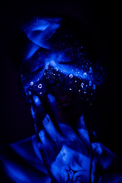 Alessia stands in a front three-quarter view with her head tilted downward. Her face is covered in an abstract pattern of glowing dots and ring shapes over dark blue UV bodypaint. Bodypaint by Geli, Charlotte Bobineau, Axell and Sage Brewer. Photographed by Elad Itzkin Photography under UV light in his studio in Gesundbrunnen, Berlin.