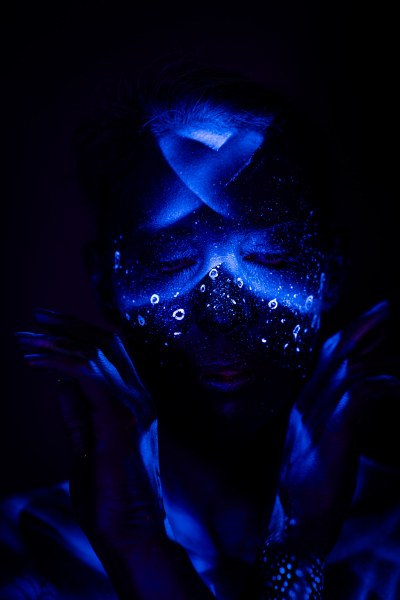 In this front-facing shot, Alessia's head is lowered. A dense pattern of glowing dots and small rings covers her face and forehead in blue UV bodypaint. Bodypaint by Geli, Charlotte Bobineau, Axell and Sage Brewer. Photographed by Elad Itzkin Photography under UV light in his Gesundbrunnen, Berlin studio.