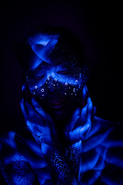 Alessia is shown standing, facing the camera with her head tilted down. Her face and forehead are painted with an abstract pattern of sparkling dots and rings that glows blue under UV light. Bodypaint by Geli, Charlotte Bobineau, Axell and Sage Brewer. Photographed by Elad Itzkin Photography in his studio in Gesundbrunnen, Berlin.