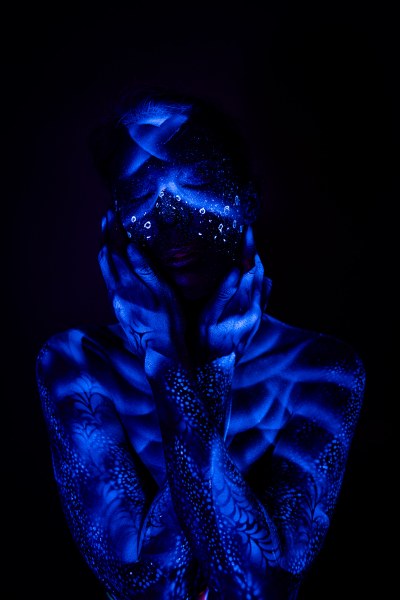 Alessia stands facing forward with her head lowered slightly, her shoulders and collarbone visible. An abstract dotted and ringed pattern in glowing blue covers her face. Bodypaint by Geli, Charlotte Bobineau, Axell and Sage Brewer. Photographed by Elad Itzkin Photography under UV light in his Gesundbrunnen, Berlin studio.