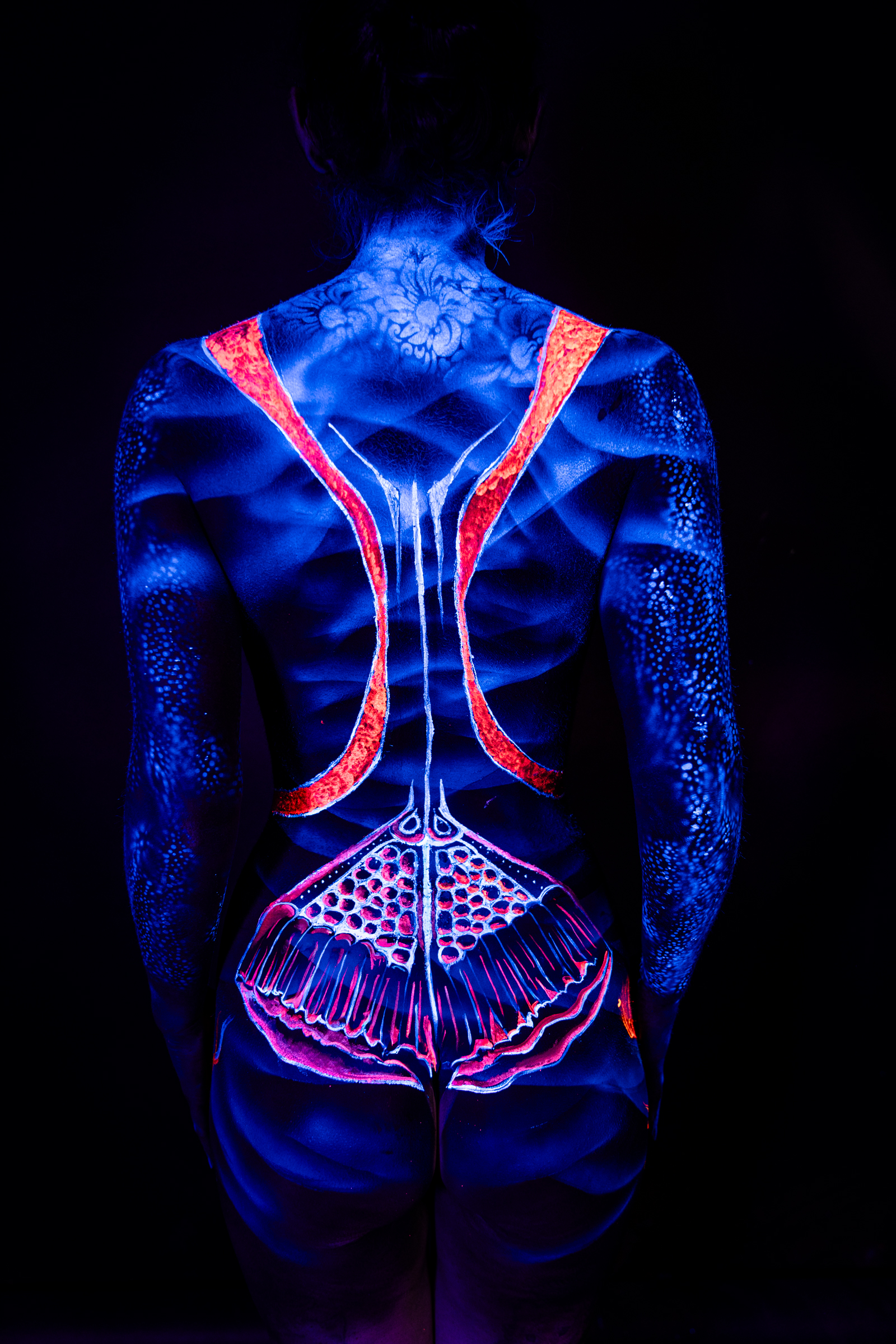 Alessia stands with her back turned to the camera. A symmetrical design of red and orange straps runs down her spine, ending in a fan-shaped mandala pattern at the lower back, with a small floral motif near the shoulders, all against blue UV-lit skin. Bodypaint by Geli, Charlotte Bobineau, Axell and Sage Brewer. Photographed by Elad Itzkin Photography in his Gesundbrunnen, Berlin studio.