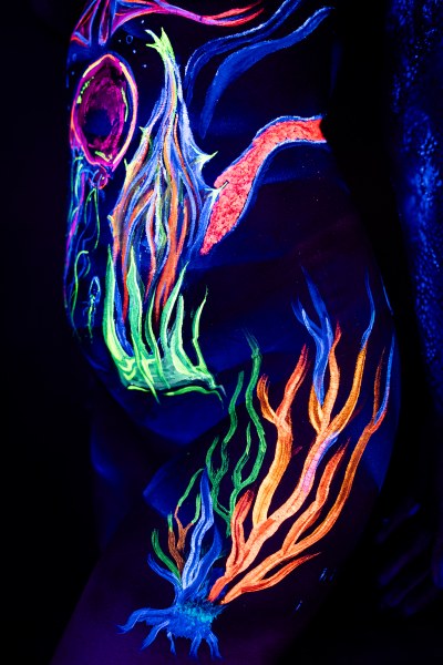 This close-up shows Alessia standing, framing her torso and hip. An abstract flame-like design in green, orange, and blue winds across her side against dark blue UV bodypaint. Bodypaint by Geli, Charlotte Bobineau, Axell and Sage Brewer. Photographed by Elad Itzkin Photography under UV light in his Gesundbrunnen, Berlin studio.