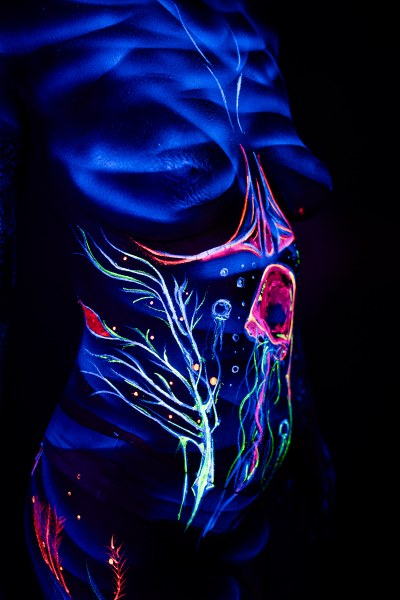 Alessia stands facing the camera in this close-up of her torso and abdomen. An abstract branching design in green and orange is paired with a round, pink jellyfish-like shape near her ribs. Bodypaint by Geli, Charlotte Bobineau, Axell and Sage Brewer. Photographed by Elad Itzkin Photography under UV light in his studio in Gesundbrunnen, Berlin.