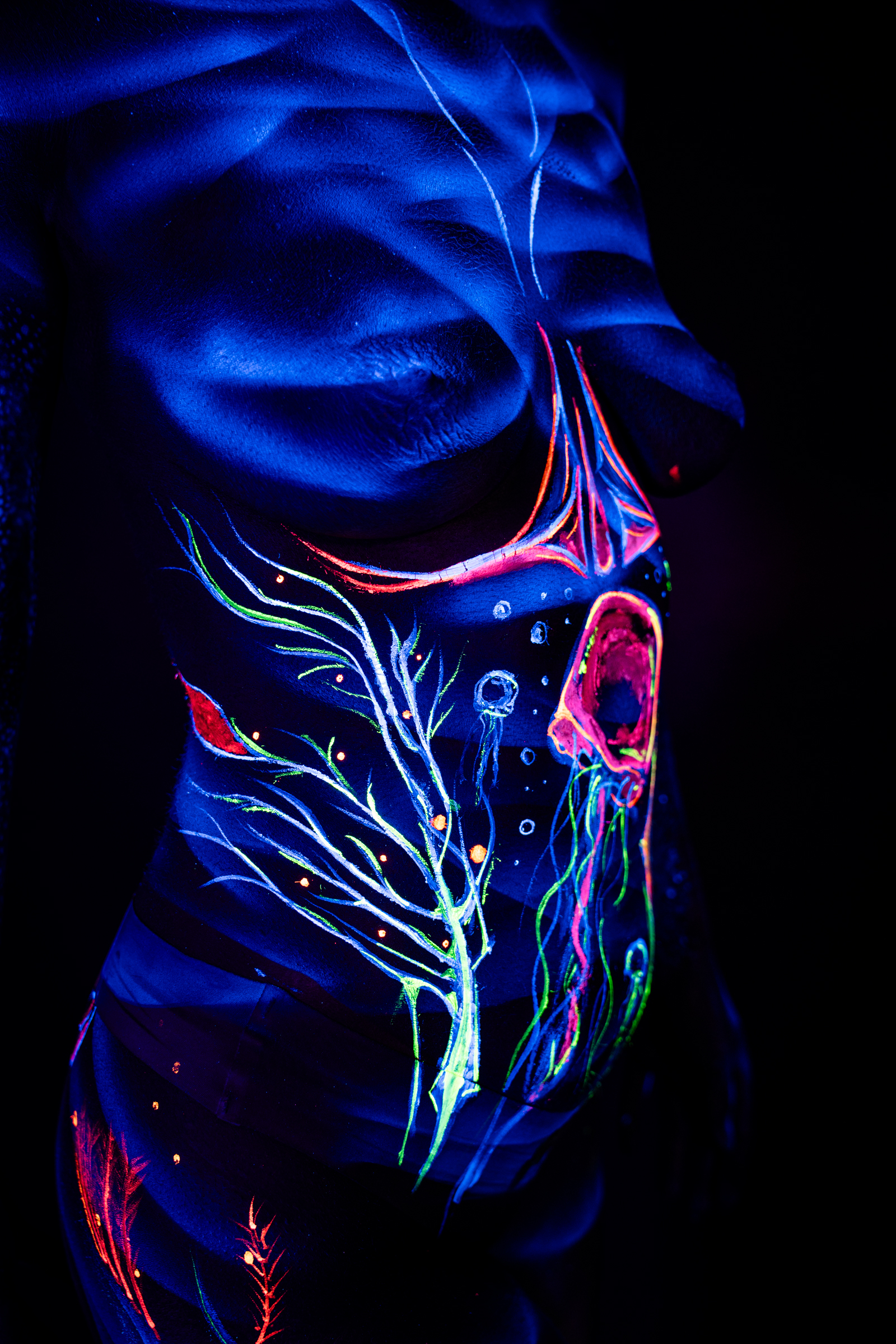 Alessia stands facing the camera in this close-up of her torso and abdomen. An abstract branching design in green and orange is paired with a round, pink jellyfish-like shape near her ribs. Bodypaint by Geli, Charlotte Bobineau, Axell and Sage Brewer. Photographed by Elad Itzkin Photography under UV light in his studio in Gesundbrunnen, Berlin.