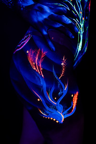 This close-up frames Alessia's hip and thigh as she stands facing the camera. An abstract feather-like design in orange and red, dotted with small circular accents, spreads across the skin under UV light. Bodypaint by Geli, Charlotte Bobineau, Axell and Sage Brewer. Photographed by Elad Itzkin Photography in his studio in Gesundbrunnen, Berlin.