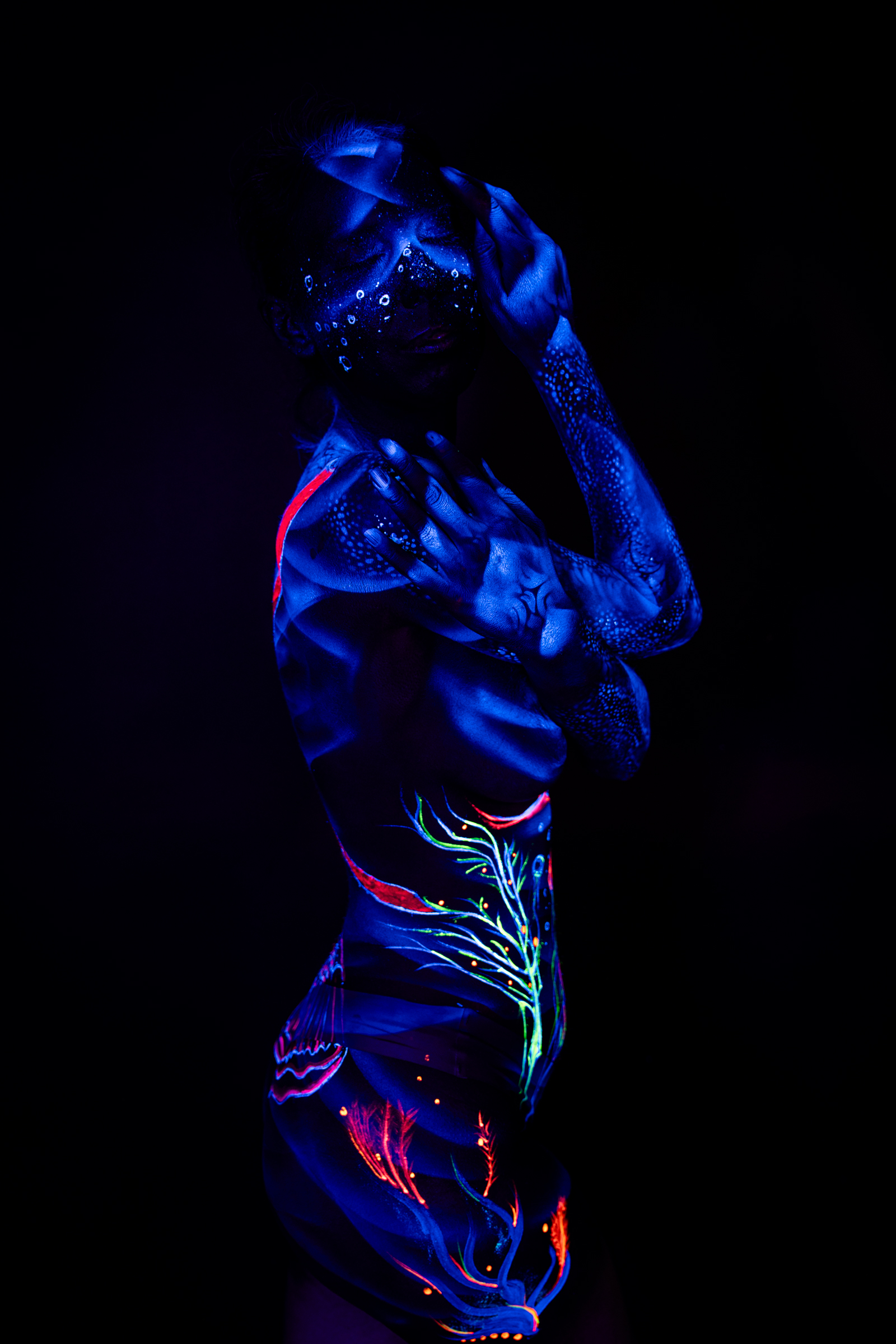 Alessia stands in profile, facing to the side. Her face carries a sparkling dotted pattern, while an abstract branching design in teal, red, and green extends down her torso under UV light. Bodypaint by Geli, Charlotte Bobineau, Axell and Sage Brewer. Photographed by Elad Itzkin Photography in his Gesundbrunnen, Berlin studio.
