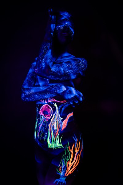 Alessia stands facing the camera in a full torso view. Her face is covered in a sparkling dotted pattern, and her abdomen carries an abstract branching design with a round pink shape, in green, orange, and pink. Bodypaint by Geli, Charlotte Bobineau, Axell and Sage Brewer. Photographed by Elad Itzkin Photography under UV light in his Gesundbrunnen, Berlin studio.
