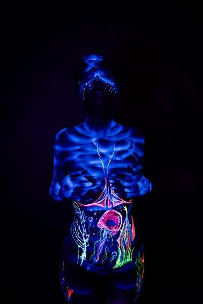 In this front-facing standing pose, Alessia's face is covered with a glowing dotted pattern. Her abdomen features an abstract branching and round pink design in green and orange tones. Bodypaint by Geli, Charlotte Bobineau, Axell and Sage Brewer. Photographed by Elad Itzkin Photography under UV light in his studio in Gesundbrunnen, Berlin.