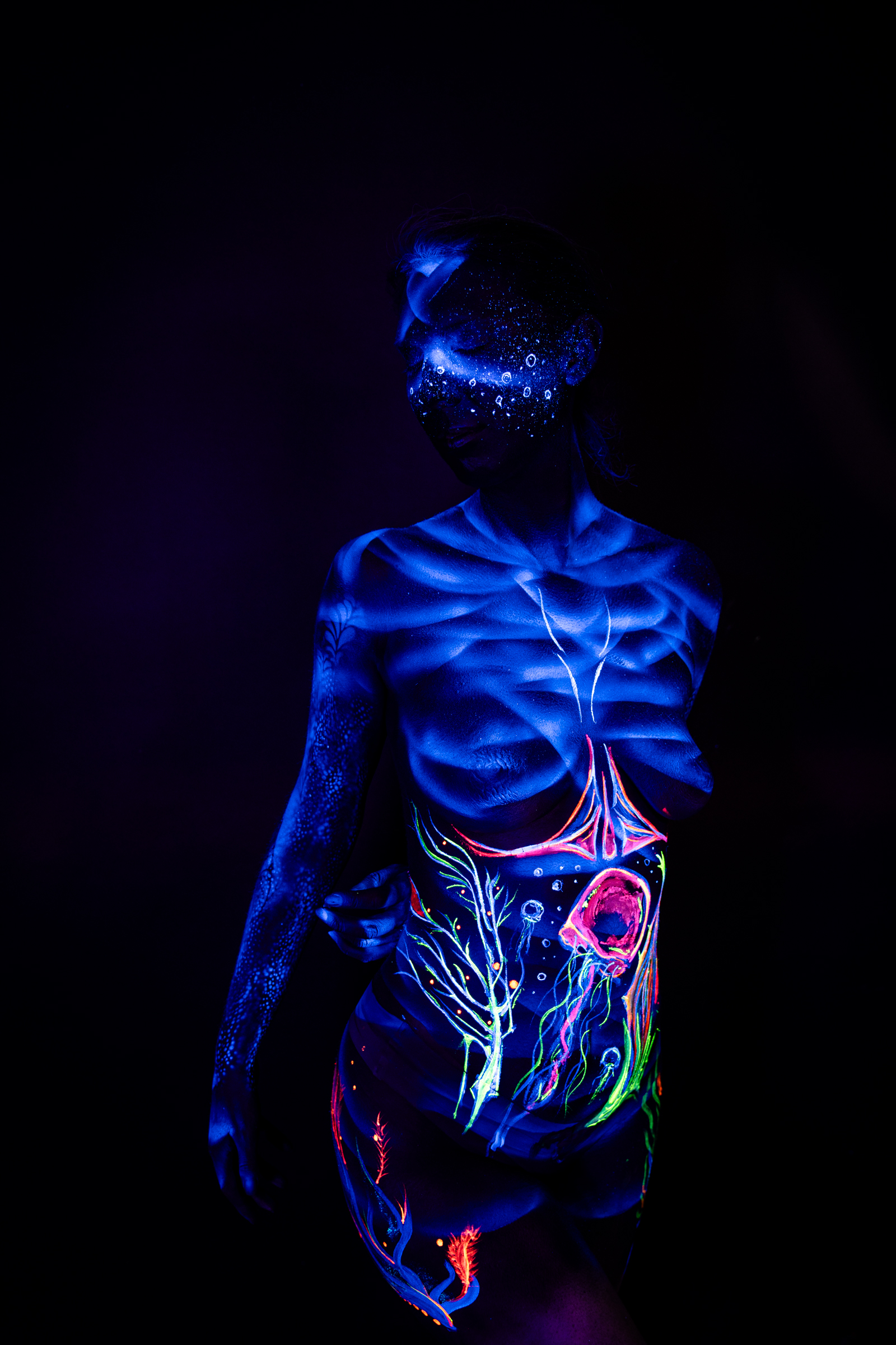 Alessia stands facing forward, her torso fully visible. An abstract branching pattern in green and orange spreads across her abdomen alongside a round pink shape, beneath a sparkling dotted design on her face. Bodypaint by Geli, Charlotte Bobineau, Axell and Sage Brewer. Photographed by Elad Itzkin Photography under UV light in his Gesundbrunnen, Berlin studio.