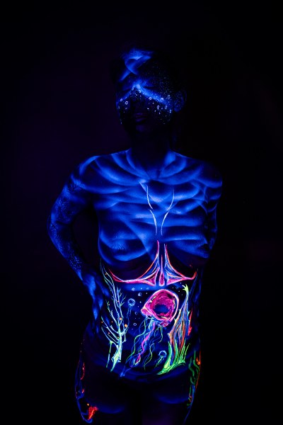 This front-view shot shows Alessia standing with her torso fully visible. Her face carries a dotted glowing pattern, and her abdomen is painted with a branching design and a round pink shape in green and orange. Bodypaint by Geli, Charlotte Bobineau, Axell and Sage Brewer. Photographed by Elad Itzkin Photography in his Gesundbrunnen, Berlin studio.