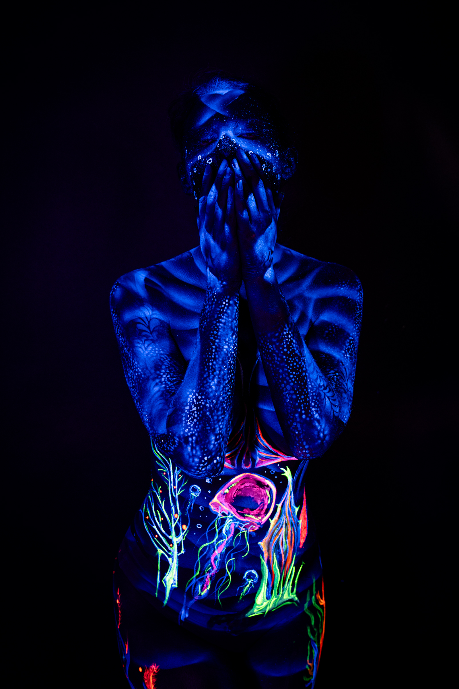Alessia stands facing forward with her head tilted down. Ribbed shading covers her torso, with a small floral accent near her shoulder and an abstract dotted pattern on her face. Bodypaint by Geli, Charlotte Bobineau, Axell and Sage Brewer. Photographed by Elad Itzkin Photography under UV light in his Gesundbrunnen, Berlin studio.