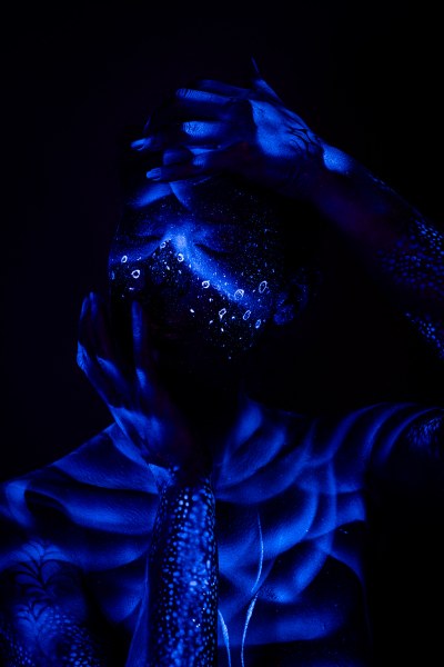This front-facing shot shows Alessia standing, her torso marked with ribbed shading and a red accent line along one side. Her face carries a sparkling dotted pattern under UV light. Bodypaint by Geli, Charlotte Bobineau, Axell and Sage Brewer. Photographed by Elad Itzkin Photography in his studio in Gesundbrunnen, Berlin.