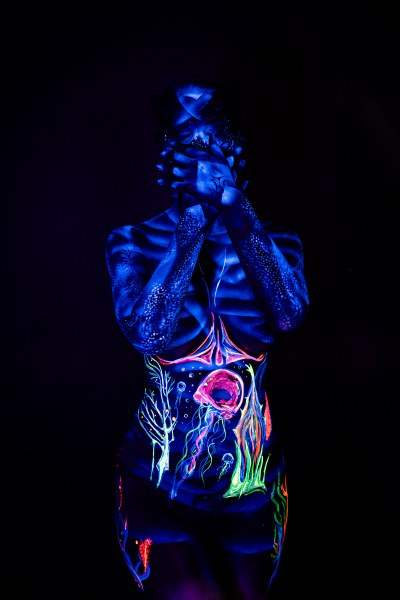 Alessia stands facing forward with her head lowered. Ribbed shading covers her upper torso, giving way to an abstract branching design and a round pink shape in green and orange on her abdomen. Bodypaint by Geli, Charlotte Bobineau, Axell and Sage Brewer. Photographed by Elad Itzkin Photography under UV light in his studio in Gesundbrunnen, Berlin.