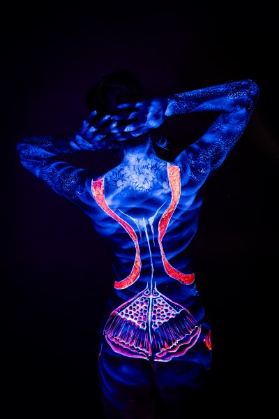 Alessia stands with her back to the camera, her head tilted back. A symmetrical design of red and orange straps runs down her spine to a fan-shaped mandala pattern at the lower back, with a floral motif near the shoulders. Bodypaint by Geli, Charlotte Bobineau, Axell and Sage Brewer. Photographed by Elad Itzkin Photography under UV light in his Gesundbrunnen, Berlin studio.