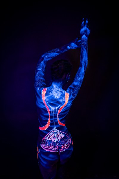 Alessia stands with her back to the camera. Her body is painted in UV blue with bright orange stripes framing the spine and a pale dotted pattern across the lower back, an abstract linework design. Bodypaint by Geli, Charlotte Bobineau, Axell, Sage Brewer. Photographed by Elad Itzkin Photography under UV light in his studio in Gesundbrunnen, Berlin.