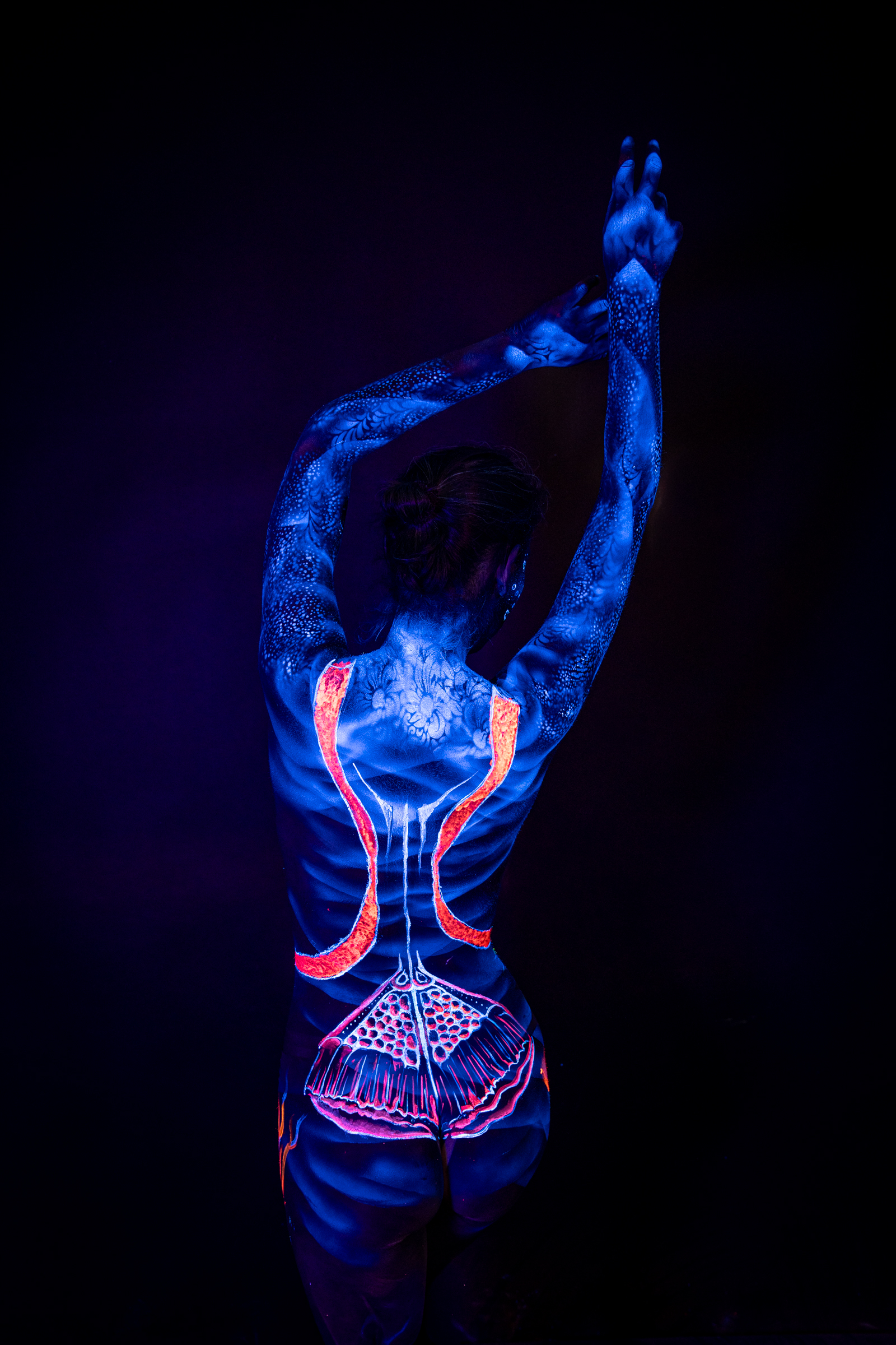 Alessia stands with her back to the camera. Her body is painted in UV blue with bright orange stripes framing the spine and a pale dotted pattern across the lower back, an abstract linework design. Bodypaint by Geli, Charlotte Bobineau, Axell, Sage Brewer. Photographed by Elad Itzkin Photography under UV light in his studio in Gesundbrunnen, Berlin.
