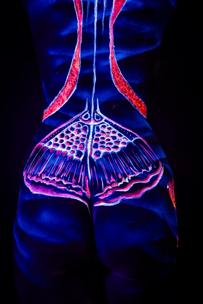 A close-up view of Alessia's lower back and hips shows an abstract UV bodypaint design in orange and pink, with a symmetrical dotted pattern forming a fan shape at the base of the spine. Bodypaint by Geli, Charlotte Bobineau, Axell, Sage Brewer. Photographed by Elad Itzkin Photography under blacklight in his studio in Gesundbrunnen, Berlin.