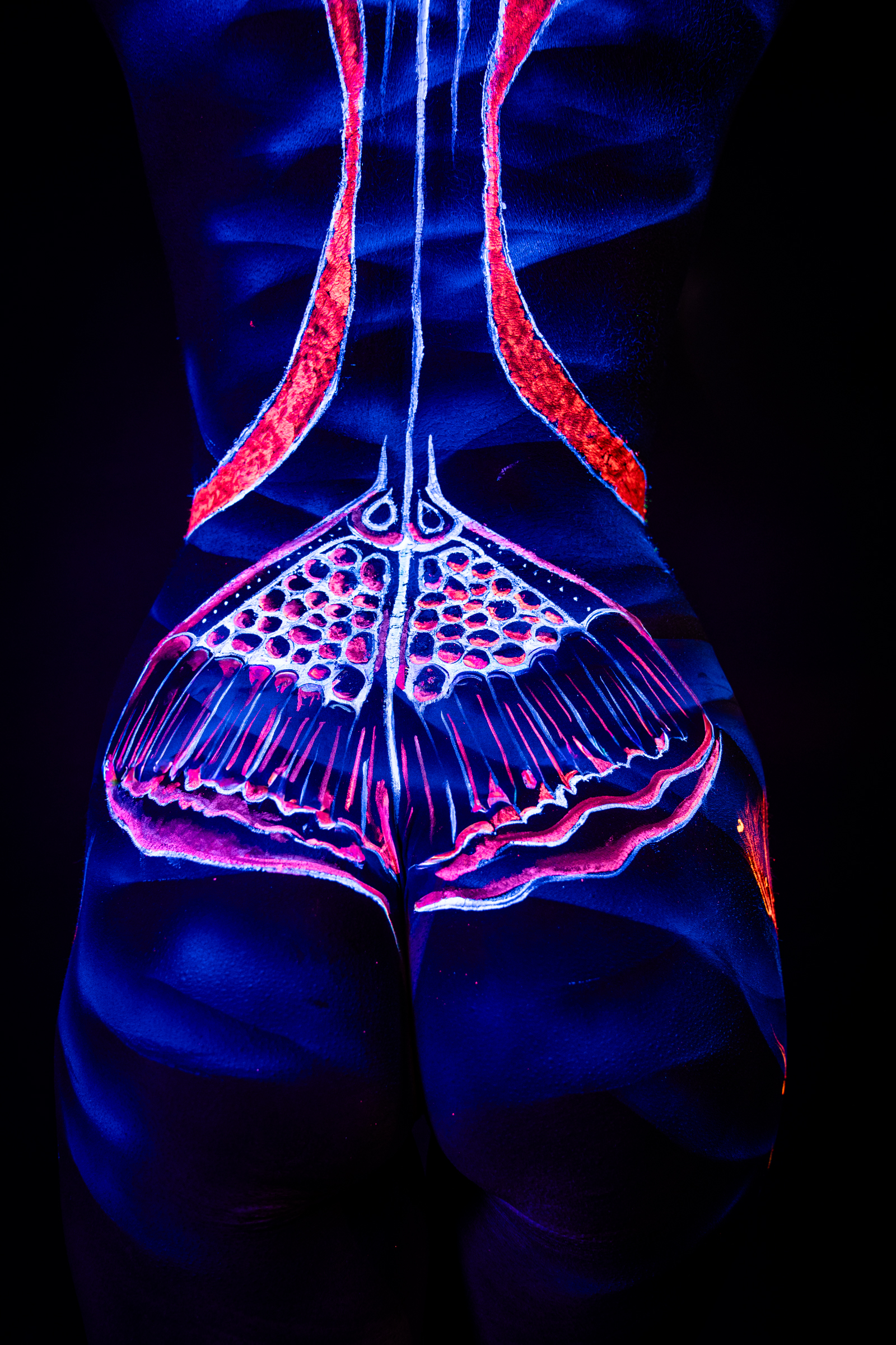 A close-up view of Alessia's lower back and hips shows an abstract UV bodypaint design in orange and pink, with a symmetrical dotted pattern forming a fan shape at the base of the spine. Bodypaint by Geli, Charlotte Bobineau, Axell, Sage Brewer. Photographed by Elad Itzkin Photography under blacklight in his studio in Gesundbrunnen, Berlin.