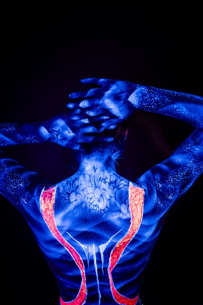 Alessia stands with her back turned to the camera, head lowered. A pale floral design covers her upper back and neck in UV paint, with bold orange stripes running down either side of the spine. Bodypaint by Geli, Charlotte Bobineau, Axell, Sage Brewer. Photographed by Elad Itzkin Photography under UV light in his studio in Gesundbrunnen, Berlin.