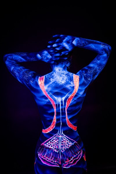 With her back to the camera and head bowed forward, Alessia is painted in glowing blue UV pigment. A soft floral pattern spans her upper back, and orange stripes trace down the spine. Bodypaint by Geli, Charlotte Bobineau, Axell, Sage Brewer. Photographed by Elad Itzkin Photography under blacklight in his studio in Gesundbrunnen, Berlin.