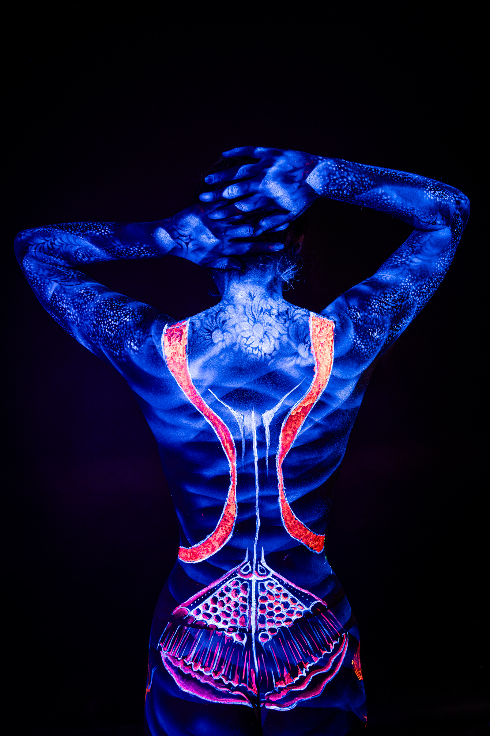 With her back to the camera and head bowed forward, Alessia is painted in glowing blue UV pigment. A soft floral pattern spans her upper back, and orange stripes trace down the spine. Bodypaint by Geli, Charlotte Bobineau, Axell, Sage Brewer. Photographed by Elad Itzkin Photography under blacklight in his studio in Gesundbrunnen, Berlin.