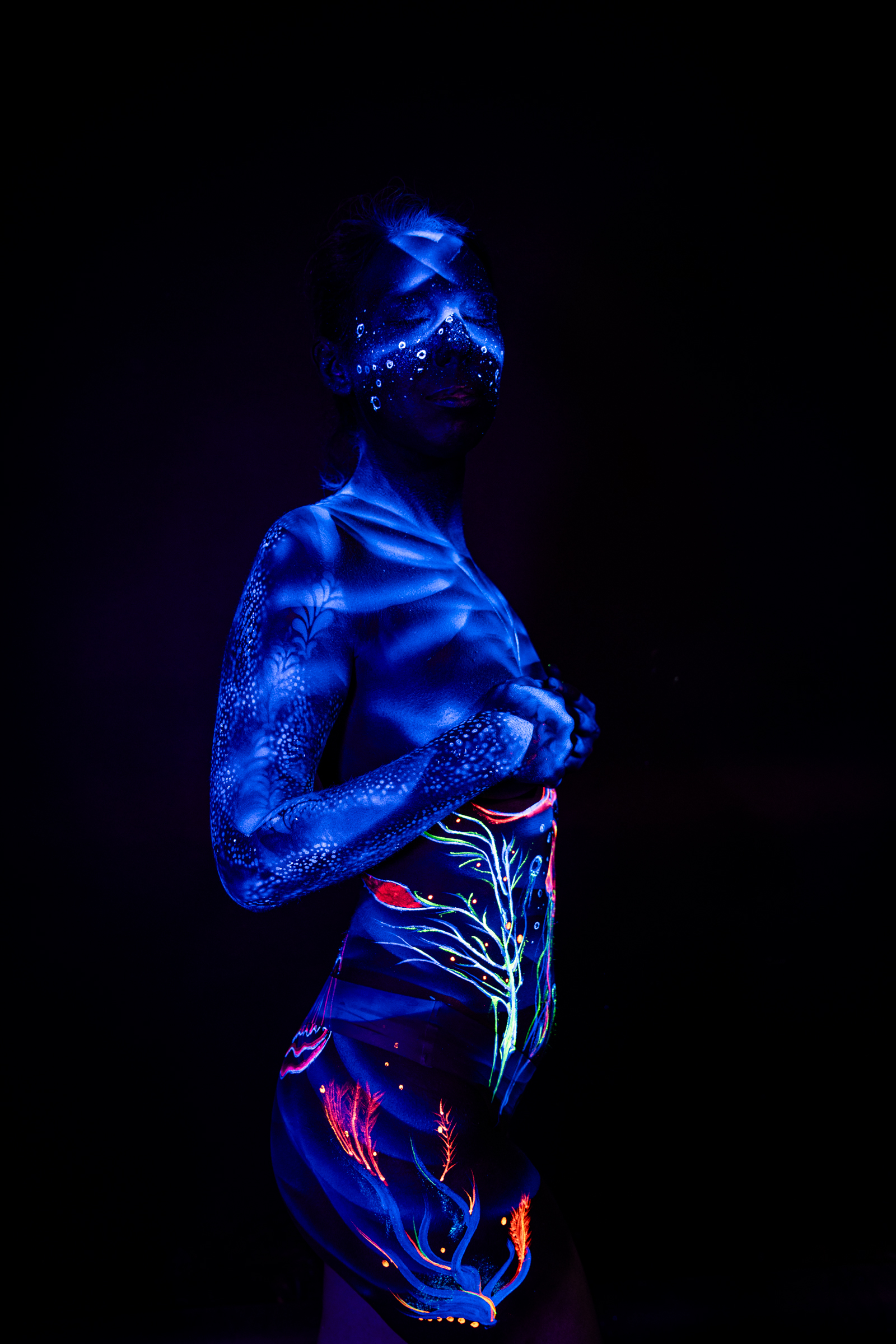 Alessia stands in a three-quarter side view. Her torso and hip are marked with a UV bodypaint design in orange and teal, featuring an abstract branch-like pattern. Bodypaint by Geli, Charlotte Bobineau, Axell, Sage Brewer. Photographed by Elad Itzkin Photography under UV light in his studio in Gesundbrunnen, Berlin.