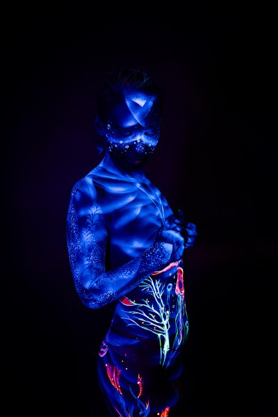 Standing in profile, Alessia's torso and hip are decorated with a UV bodypaint design in orange, teal and pink, featuring an abstract branch-like motif. Bodypaint by Geli, Charlotte Bobineau, Axell, Sage Brewer. Photographed by Elad Itzkin Photography under blacklight in his studio in Gesundbrunnen, Berlin.
