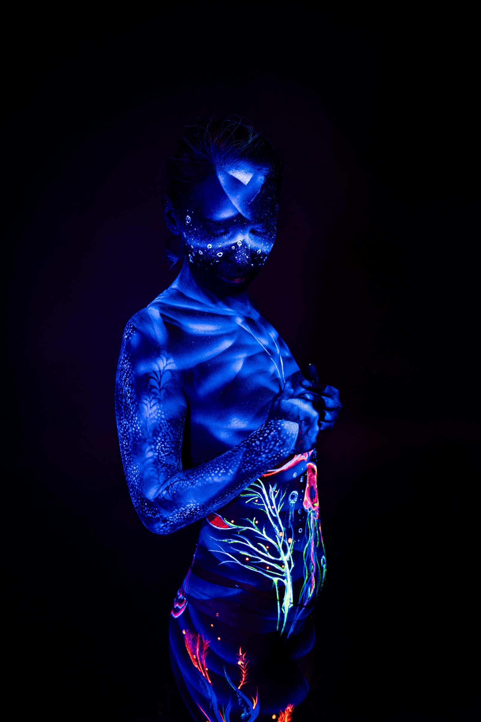 Standing in profile, Alessia's torso and hip are decorated with a UV bodypaint design in orange, teal and pink, featuring an abstract branch-like motif. Bodypaint by Geli, Charlotte Bobineau, Axell, Sage Brewer. Photographed by Elad Itzkin Photography under blacklight in his studio in Gesundbrunnen, Berlin.