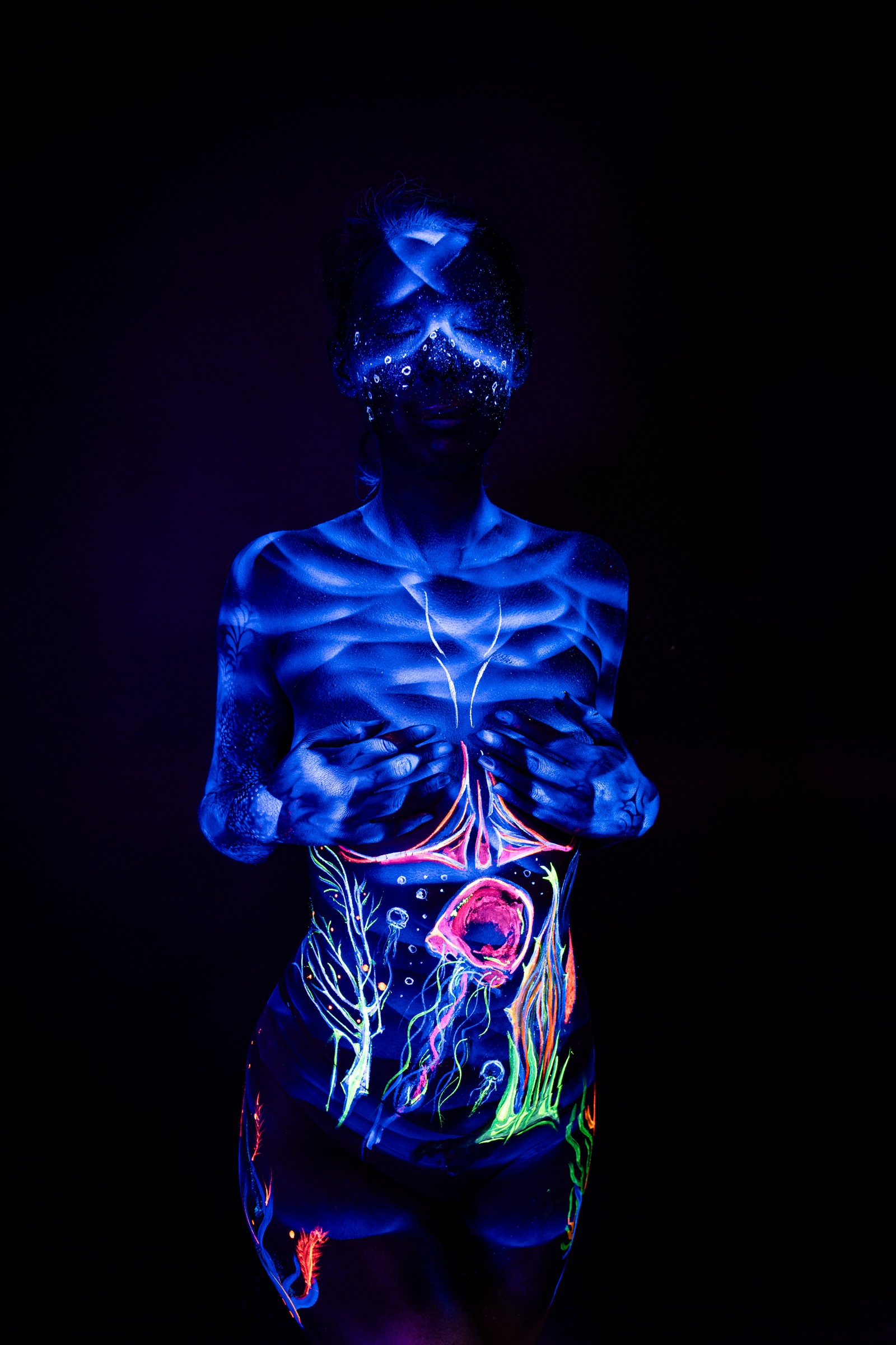 Alessia stands facing forward, her face covered in a dark UV mask pattern. Her midsection is marked with an abstract design in orange, pink and green. Bodypaint by Geli, Charlotte Bobineau, Axell, Sage Brewer. Photographed by Elad Itzkin Photography under UV light in his studio in Gesundbrunnen, Berlin.