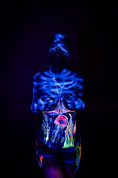 In a front-facing standing pose, Alessia wears a UV-painted mask design across her face, with a colorful abstract pattern in orange, pink and green covering her stomach. Bodypaint by Geli, Charlotte Bobineau, Axell, Sage Brewer. Photographed by Elad Itzkin Photography under blacklight in his studio in Gesundbrunnen, Berlin.