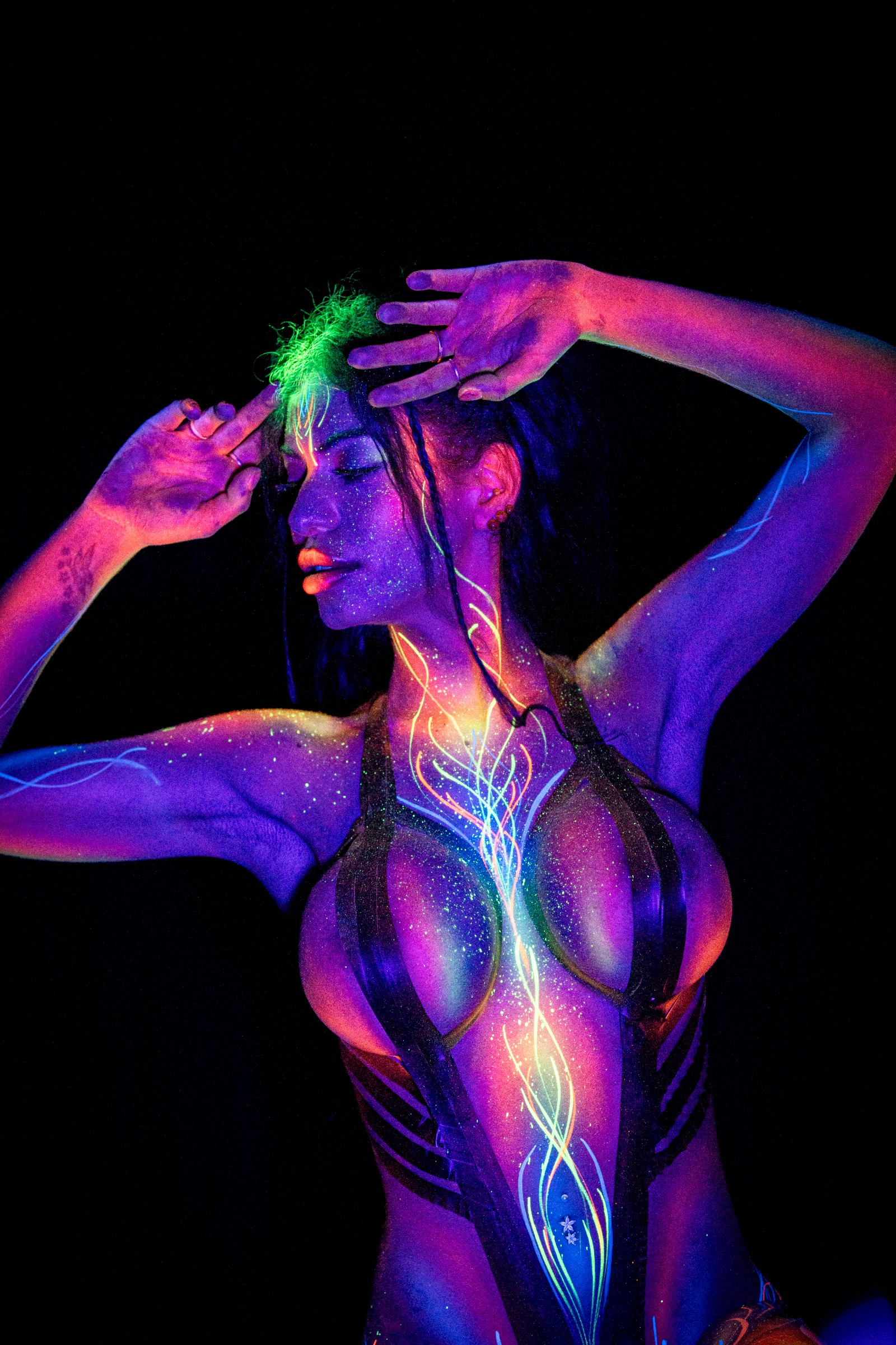 AlyssaHotofficial stands in profile with her head lowered. A glowing green and yellow linear pattern with sparkle highlights runs along one side of her torso and leg. Bodypaint by Claudia Kraemer. Photographed by Elad Itzkin Photography under UV blacklight in his Gesundbrunnen, Berlin studio.