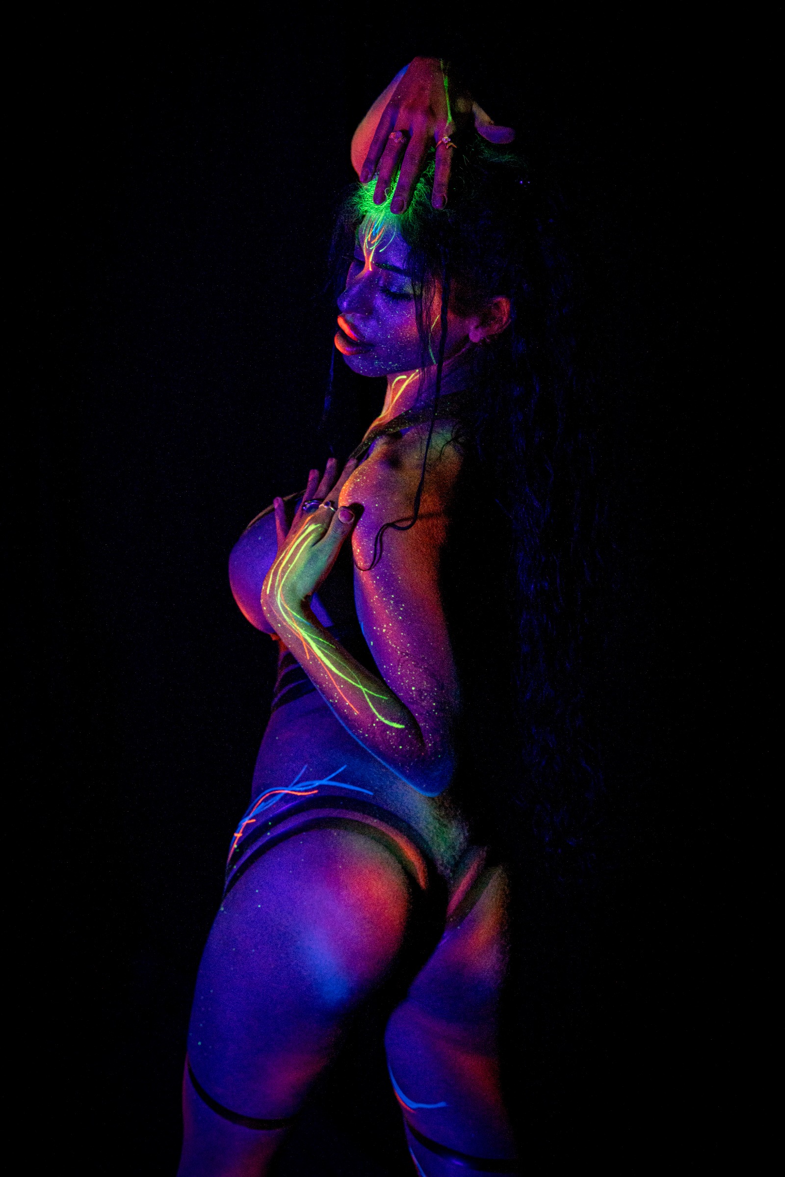 AlyssaHotofficial is shown in profile with her head tilted forward. Sparkling UV highlights and a glowing linear pattern trace across her shoulder and chest. Bodypaint by Claudia Kraemer. Photographed by Elad Itzkin Photography under UV blacklight in his studio in Gesundbrunnen, Berlin.