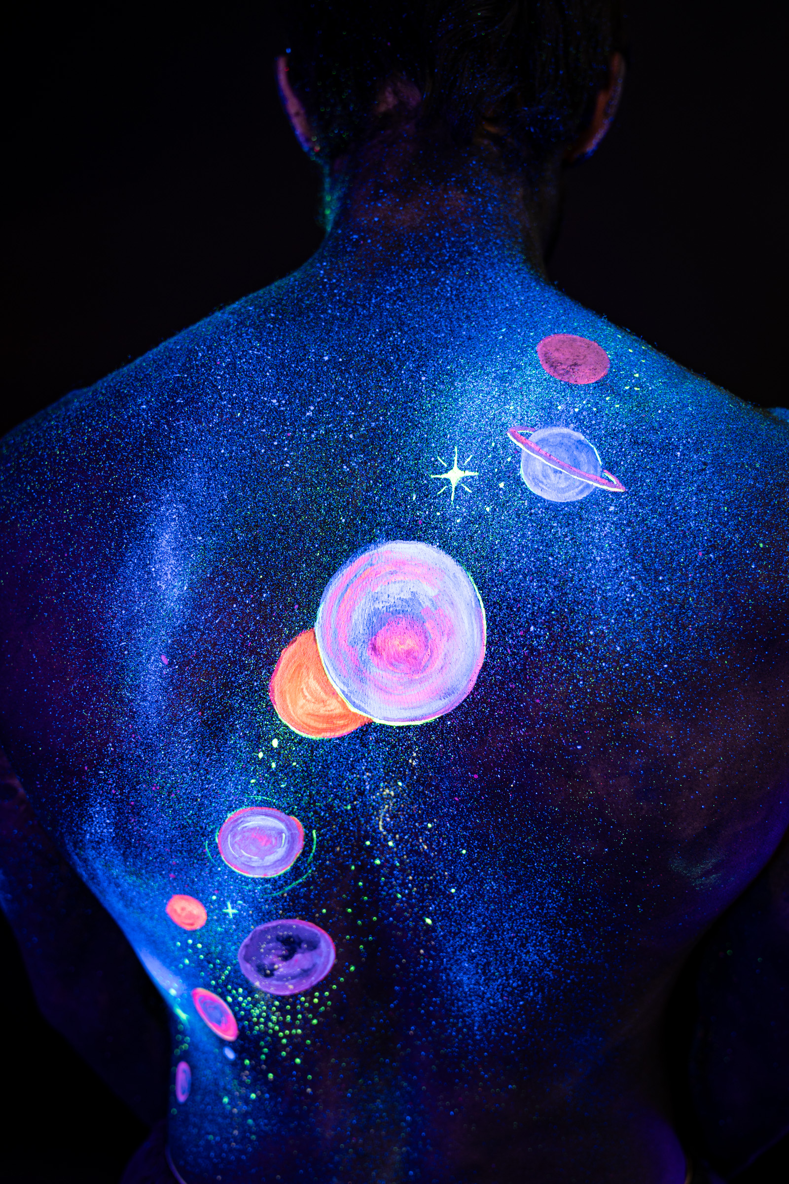 Axell, back turned to the camera. A space themed design of glowing pink, orange and blue planets covers the back over a glittering blue base. Bodypaint by Elad Itzkin, Charlotte Bobineau, Sage Brewer. Photographed by Elad Itzkin Photography under UV/blacklight in his studio in Gesundbrunnen, Berlin.